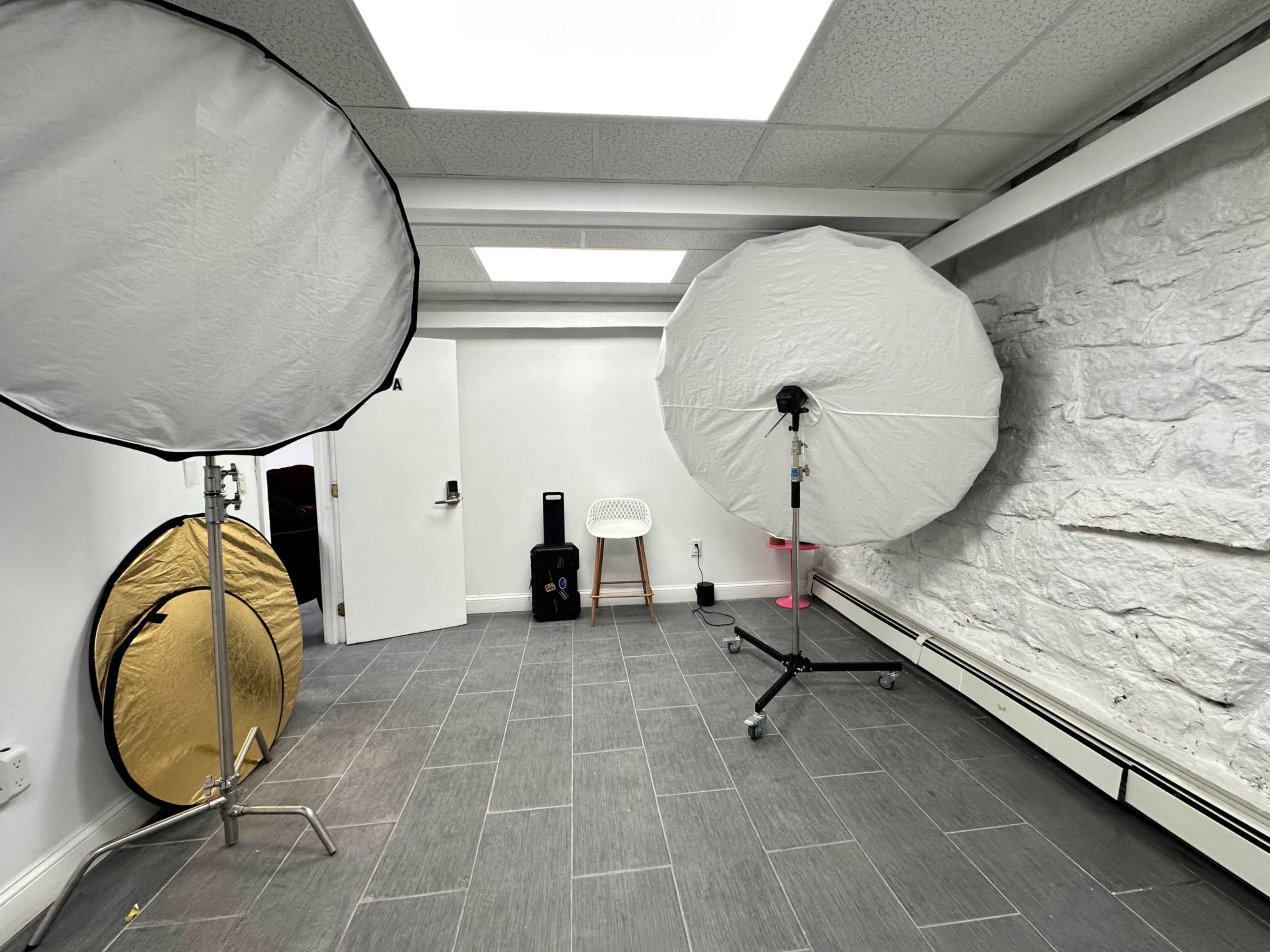 The image shows a studio room with two large softbox lights, a white chair, and a section of a stone wall.