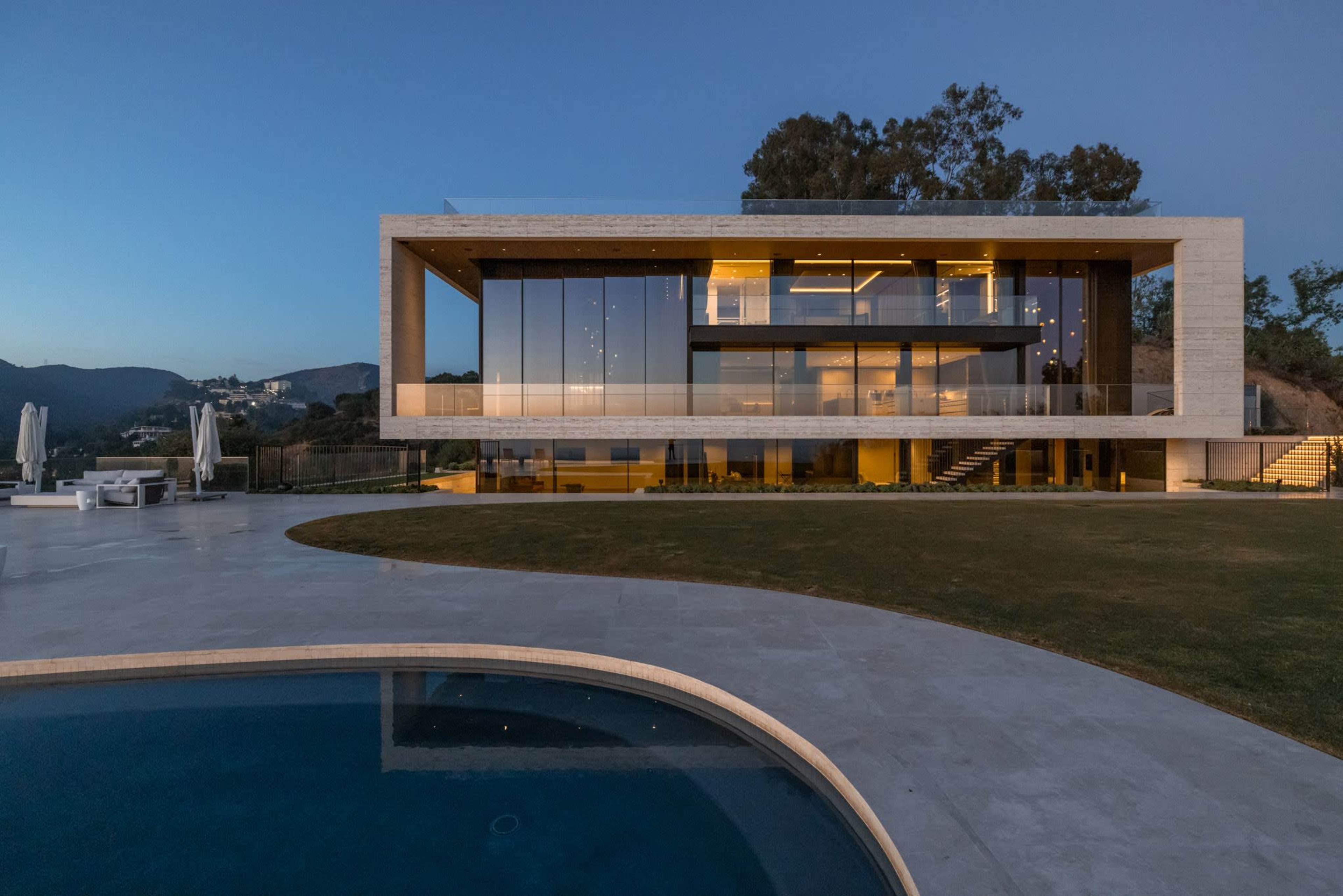A modern two-story house with large glass windows and a swimming pool is situated on a landscaped lawn at dusk.