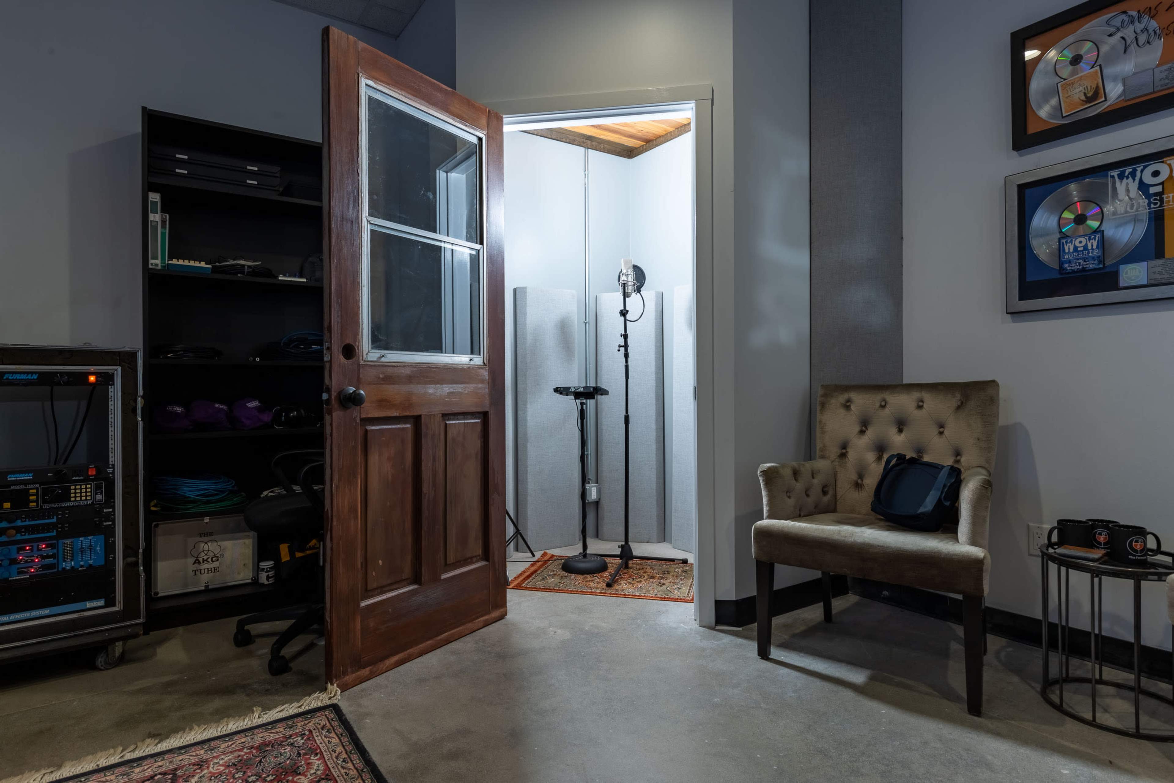 A recording studio room features an open door leading to a sound booth with a microphone on a stand, accompanied by a plush chair and equipment shelves.
