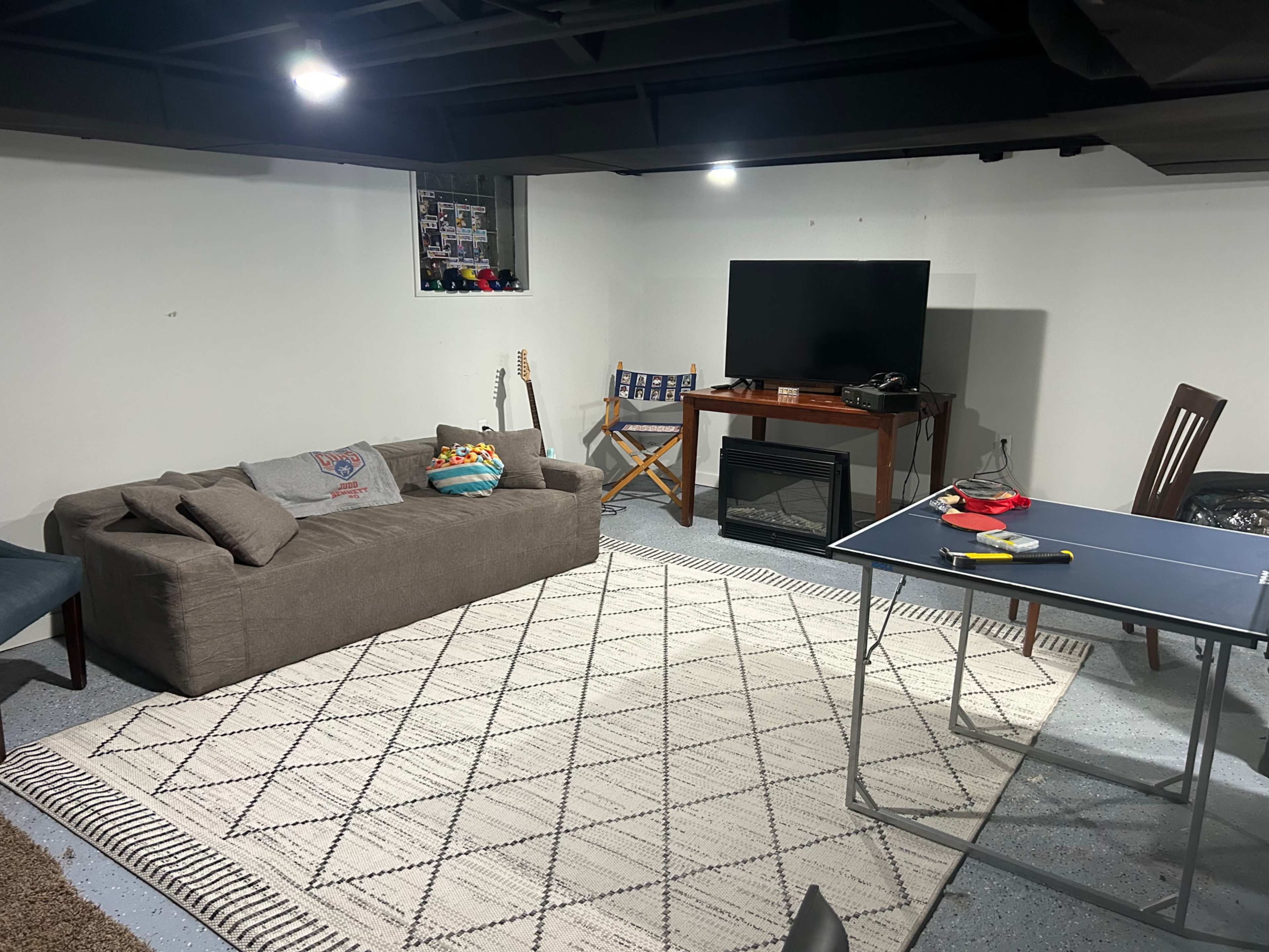 The image shows a basement living area featuring a sofa, a television on a stand, a small table with ping pong equipment, a rug, and a guitar leaning against the wall.