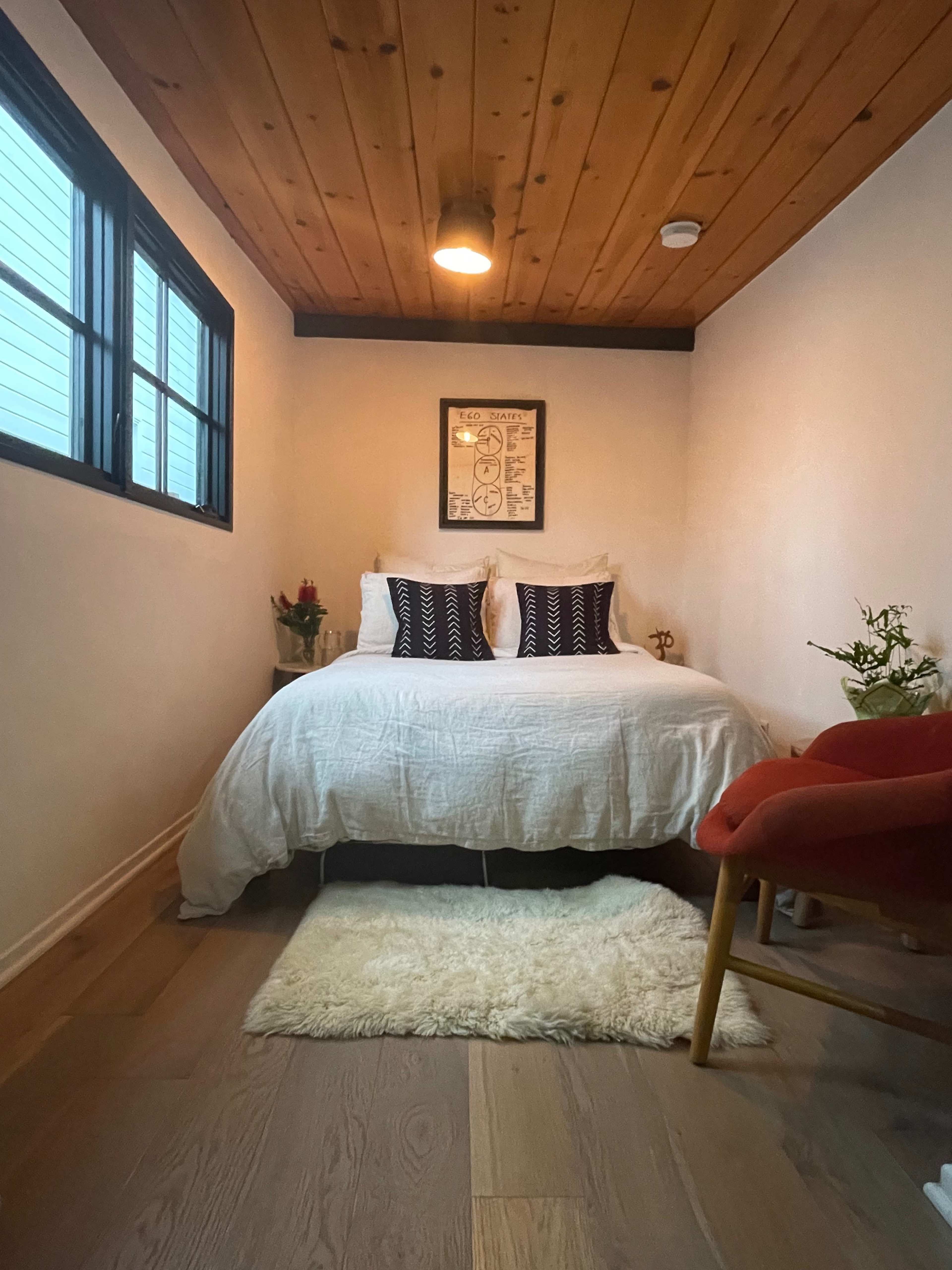 A small bedroom featuring a bed with white bedding, a wooden ceiling, a window, and a red chair beside a small rug.