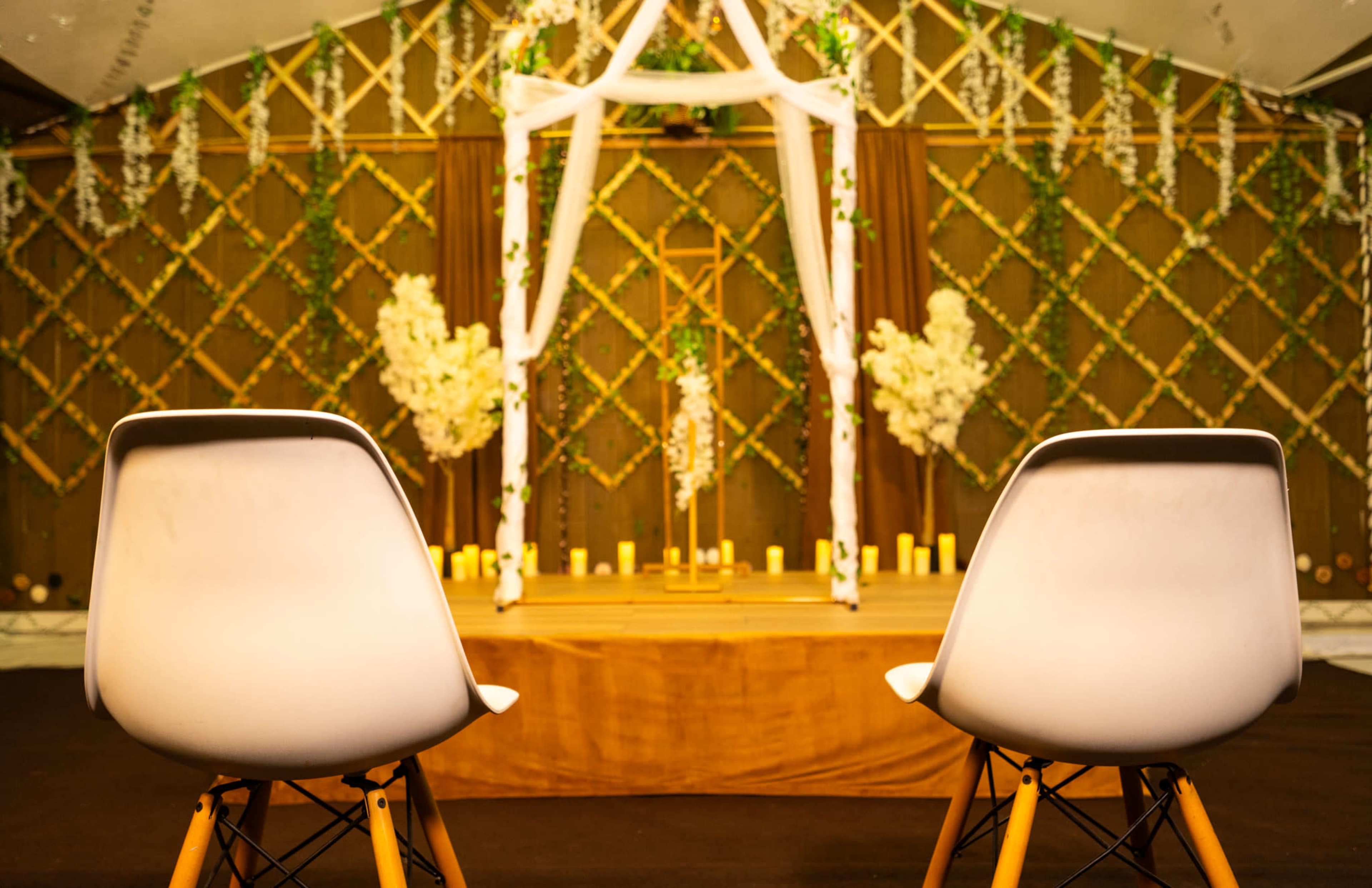 Two white chairs with wooden legs face a decorated stage featuring an arch adorned with flowers and candles in a tent setting.