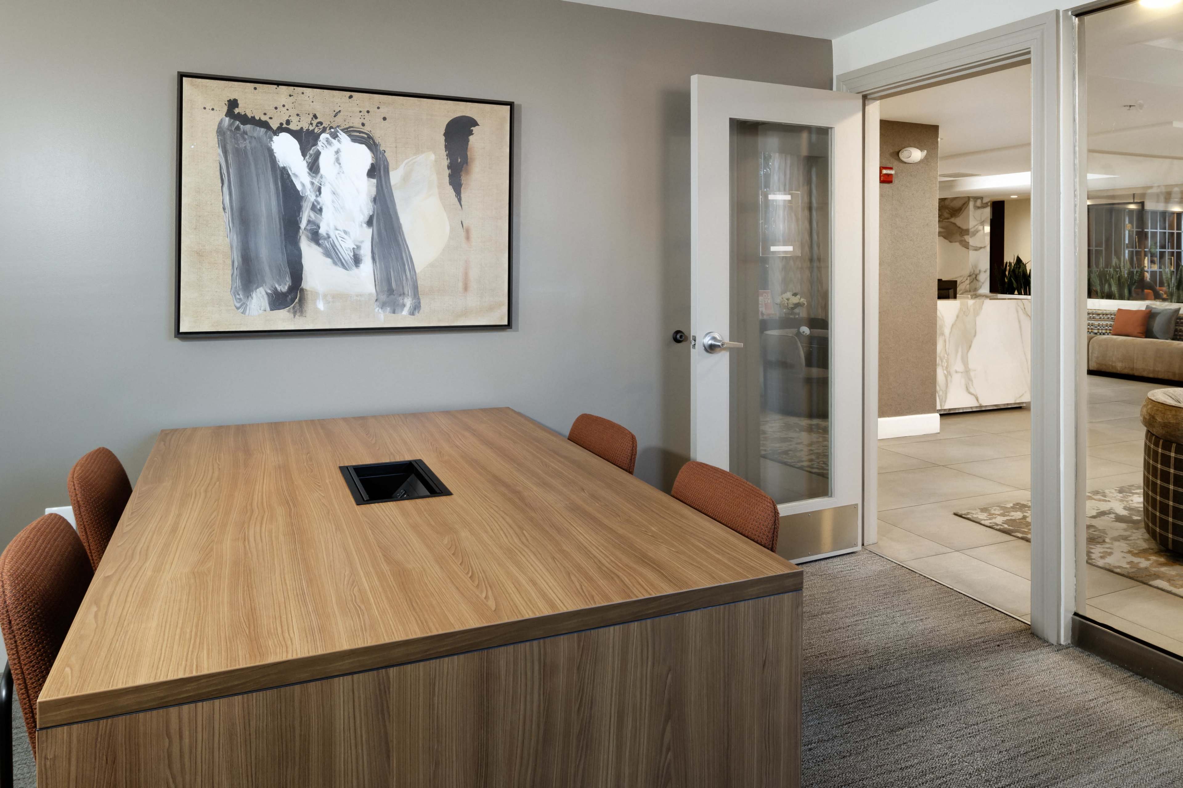 The image shows a modern conference room with a wooden table and four brown chairs, featuring an abstract artwork on the wall, and a doorway leading to another area.
