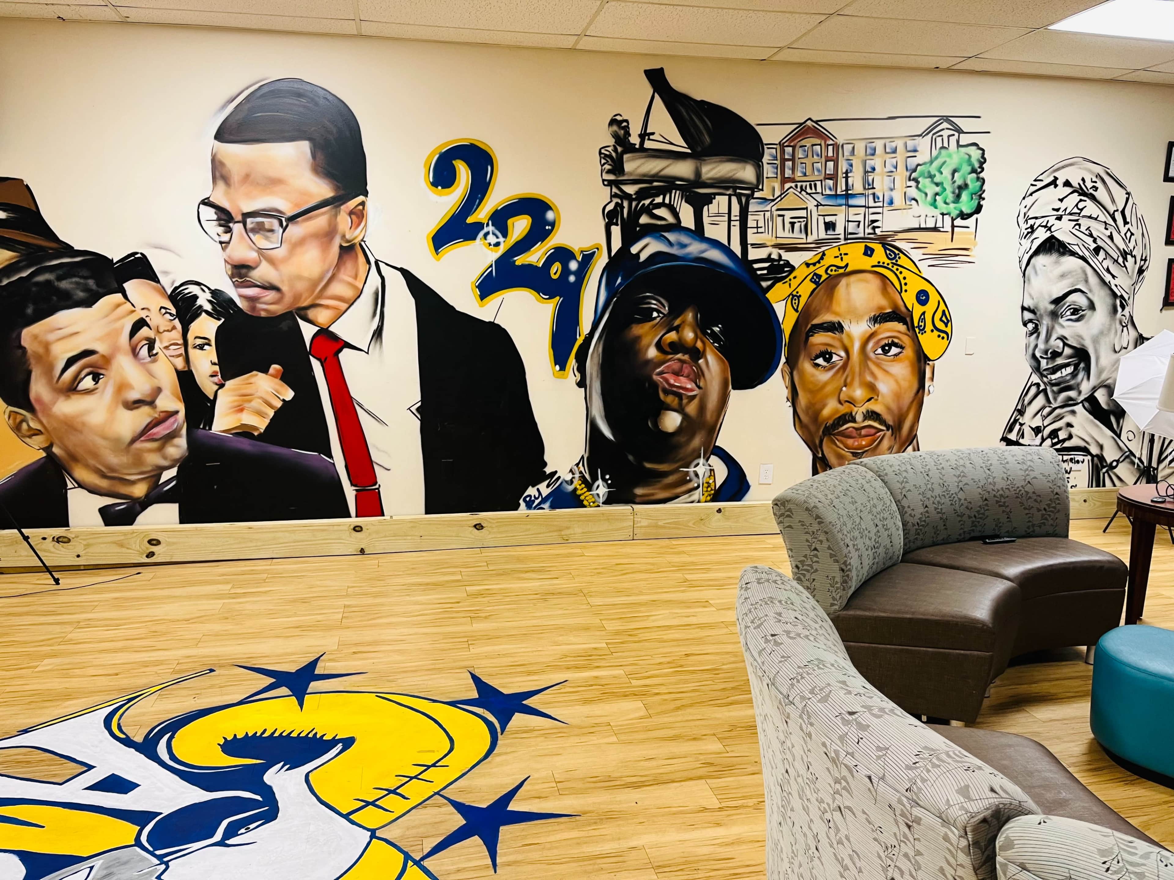 The image shows a mural featuring prominent figures from hip-hop culture along a wall in a room with a wooden floor and comfortable seating.