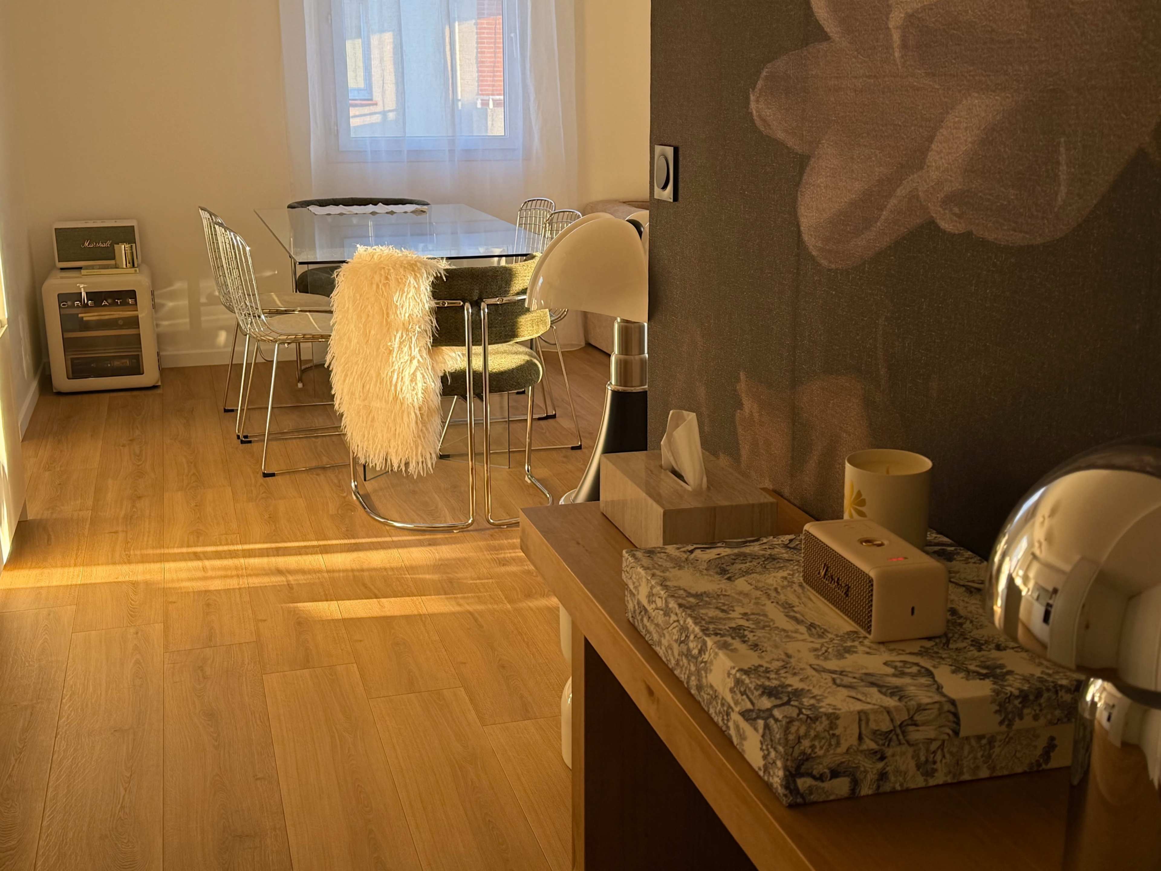 The image shows a well-lit dining area featuring a glass dining table surrounded by chrome chairs, a lamp, and a decorative box on a wooden surface.