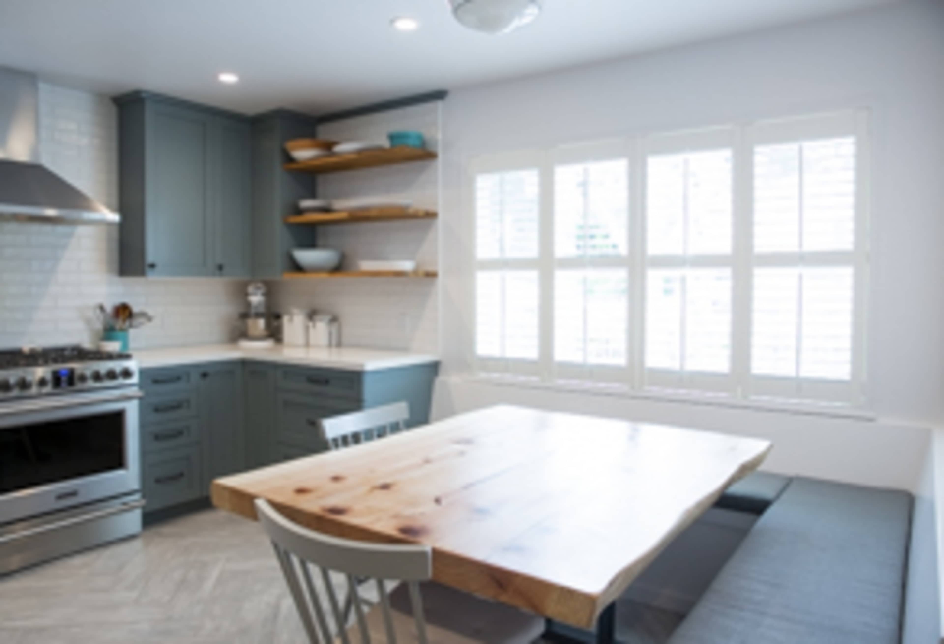 A modern kitchen features a stainless steel stove, blue cabinetry, open shelves with dishware, and a wooden dining table with grey seating next to a window.