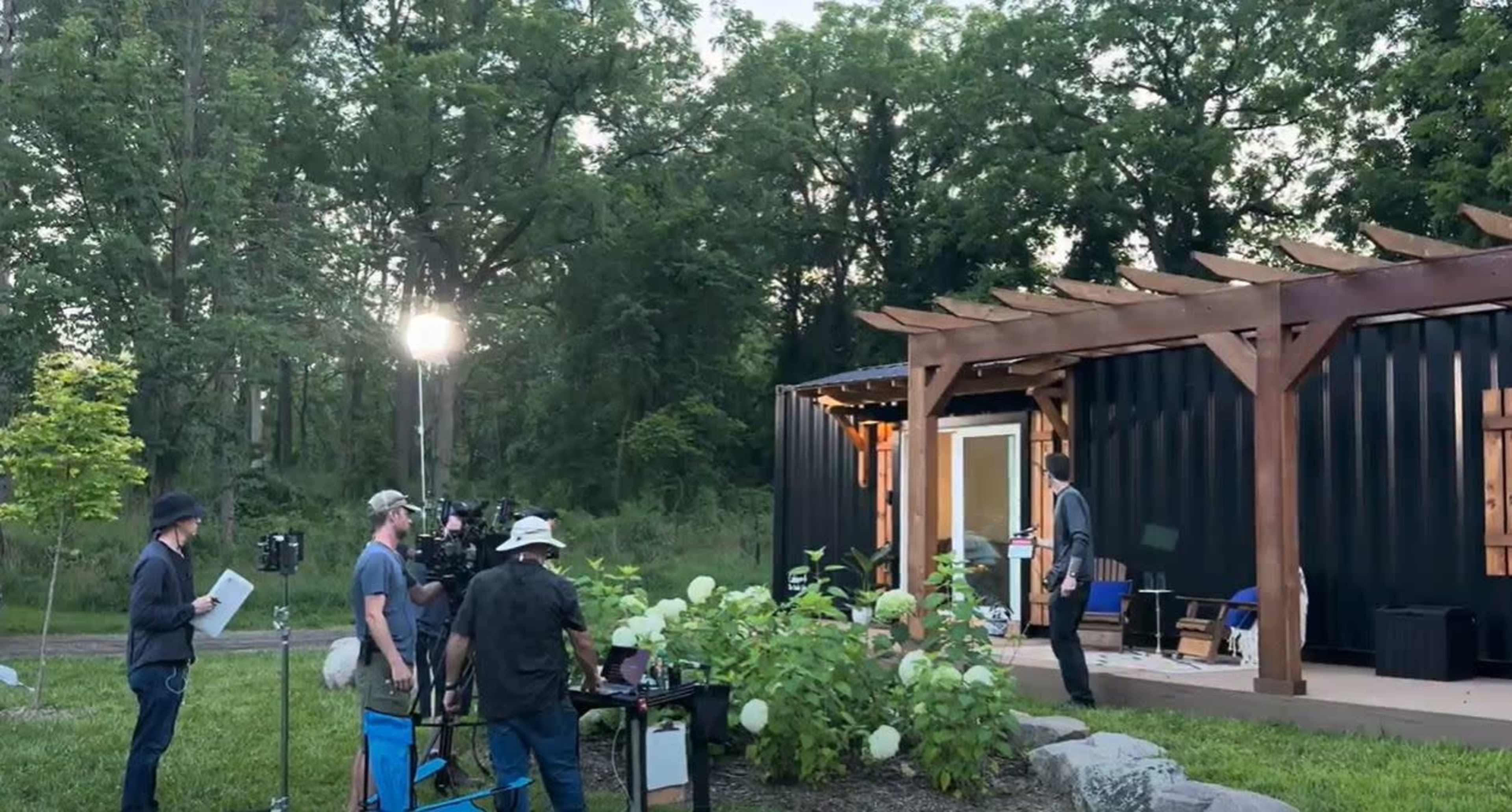 A film crew sets up cameras and equipment outside a black container-style building amidst greenery.