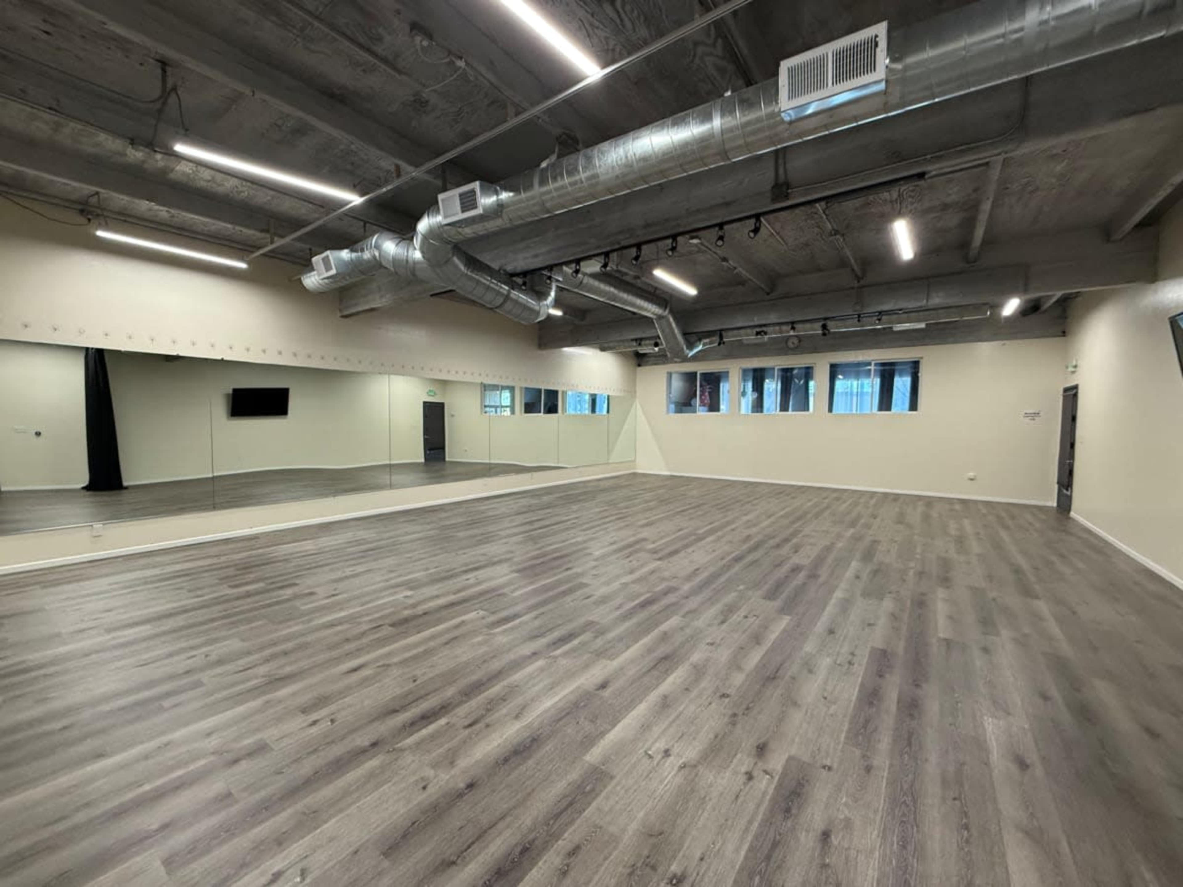 The image shows an empty dance studio with wooden flooring, large mirrors on one wall, and industrial-style ceiling ductwork.