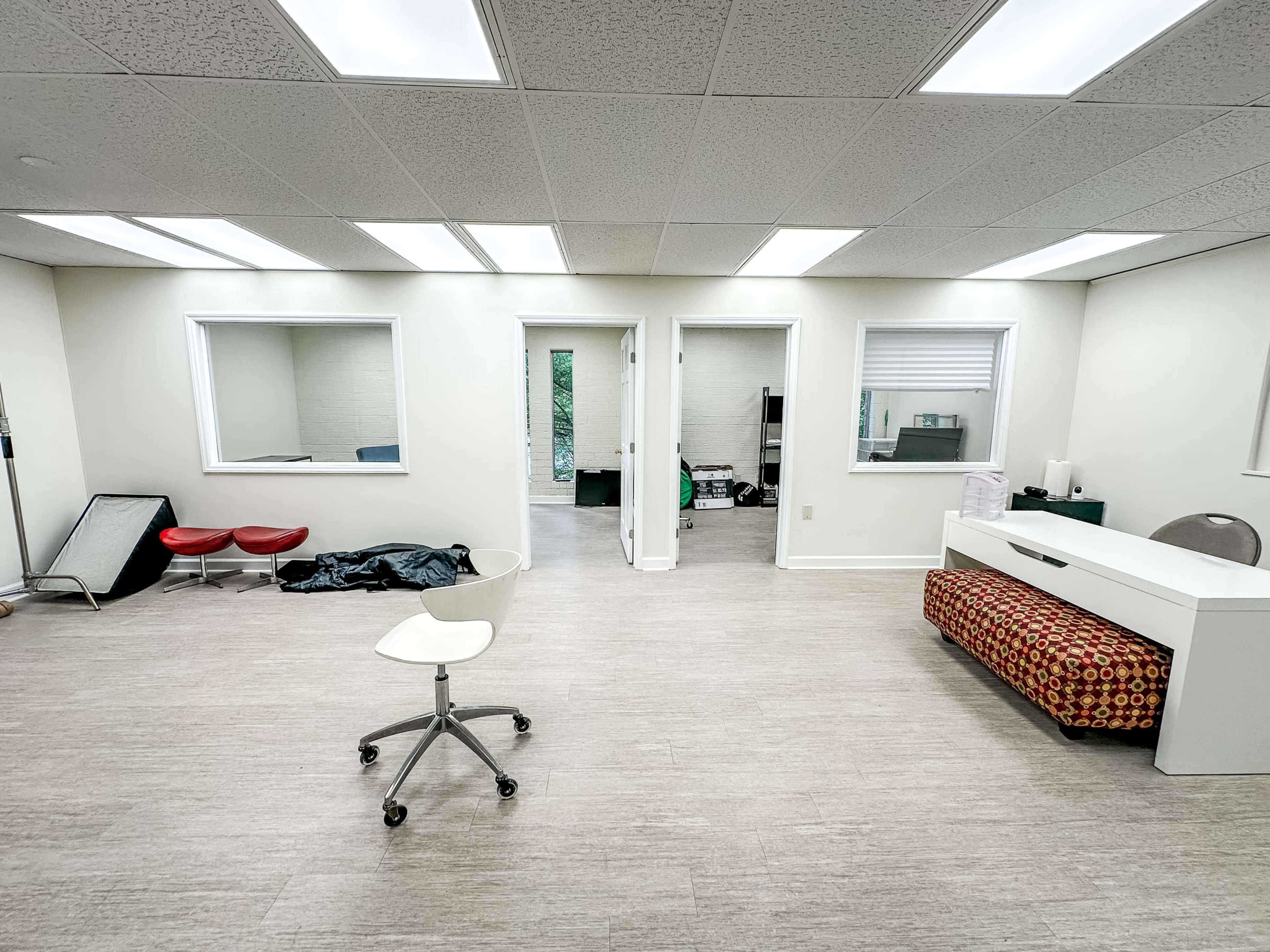 A bright, minimalistic office space with white walls, several windows, and two entrances, along with a desk and a wheeled chair.