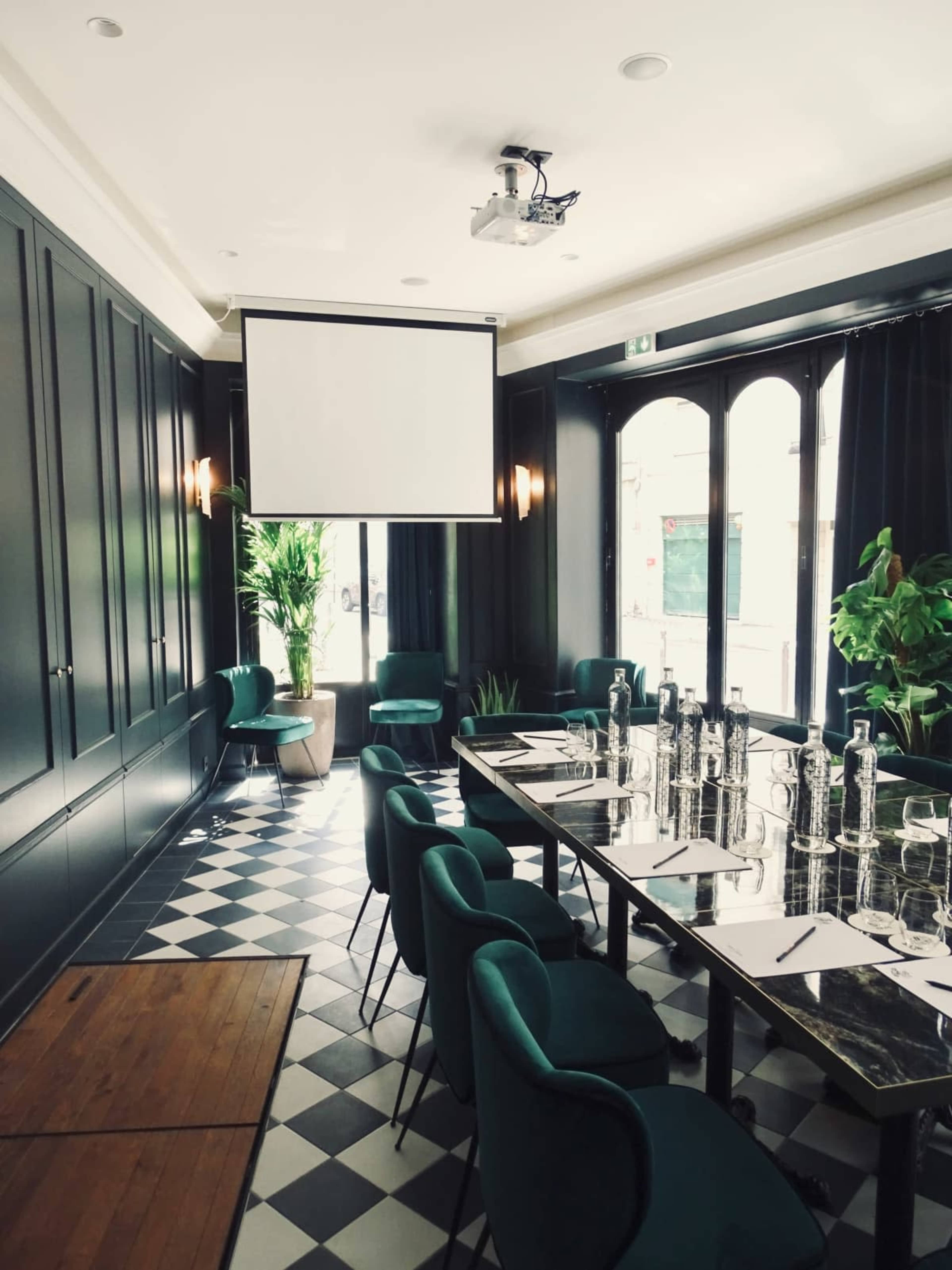 The image shows a formal dining area with dark walls, a large table set up with glassware and table settings, green upholstered chairs, and a projector screen hanging from the ceiling.