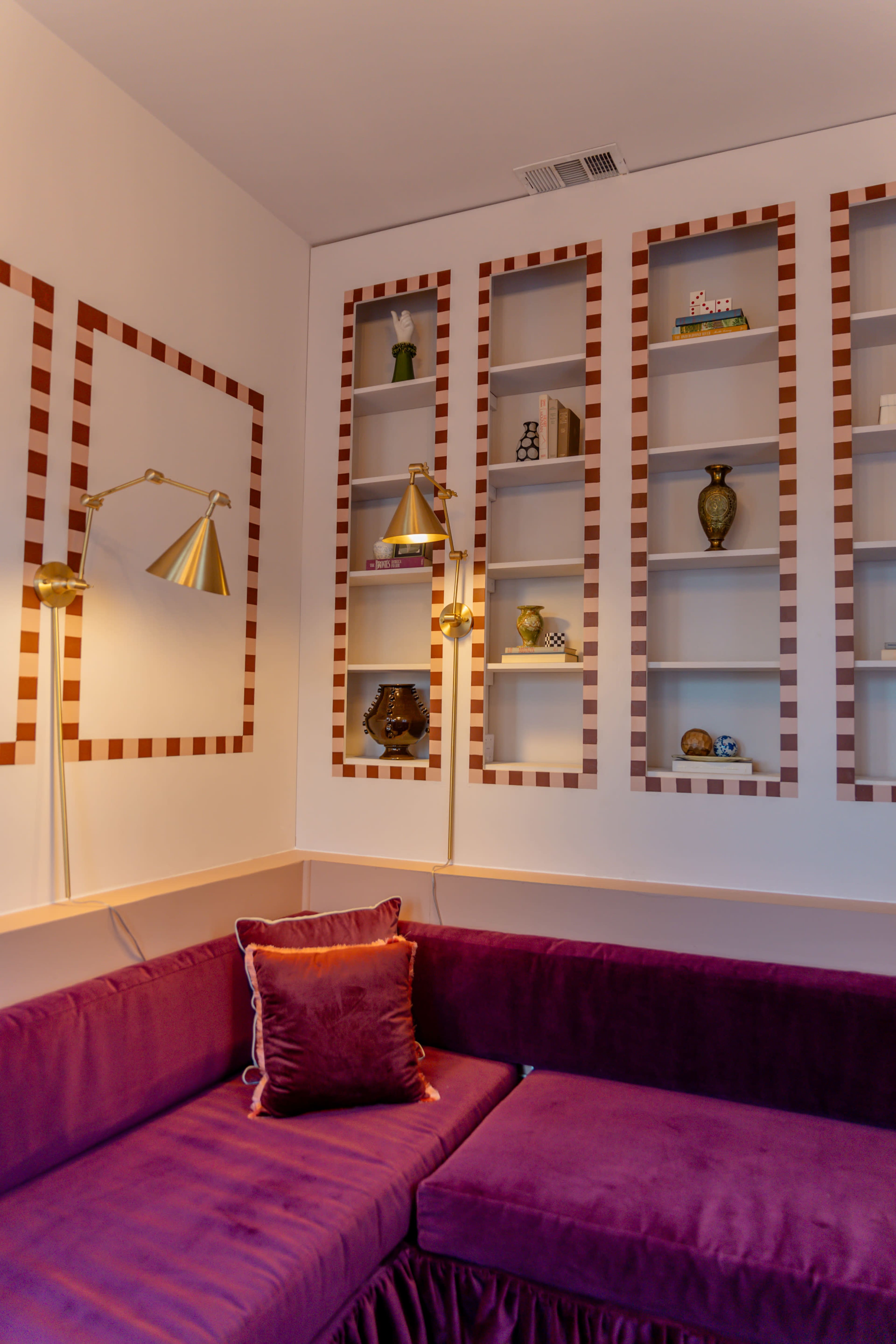 A corner of a room with a purple sofa and wall shelves displaying decorative items, illuminated by two wall-mounted lamps.