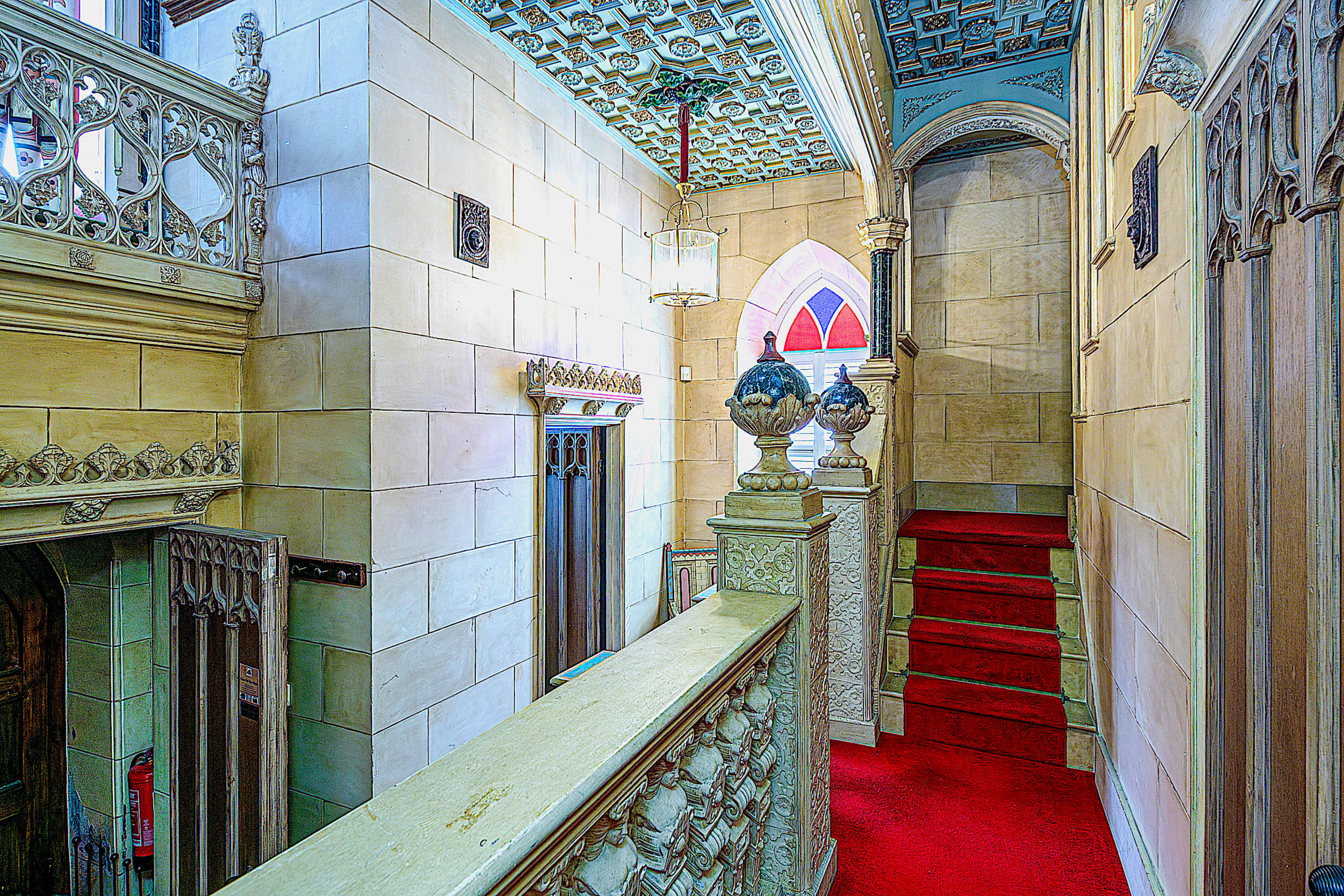 The image shows a grand staircase in an ornate hallway with intricate architecture and decorative elements, including a colorful ceiling and red carpet.