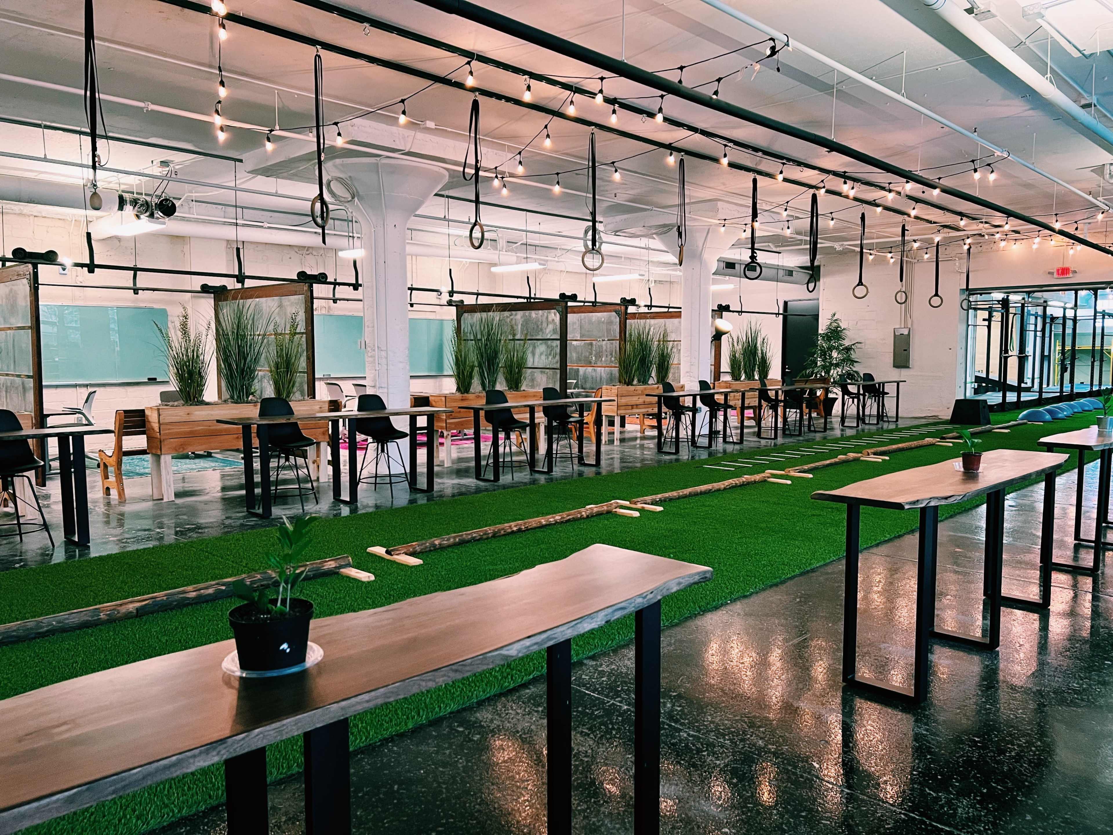 A spacious interior with rows of tables and chairs arranged on a green artificial grass surface, illuminated by string lights overhead.
