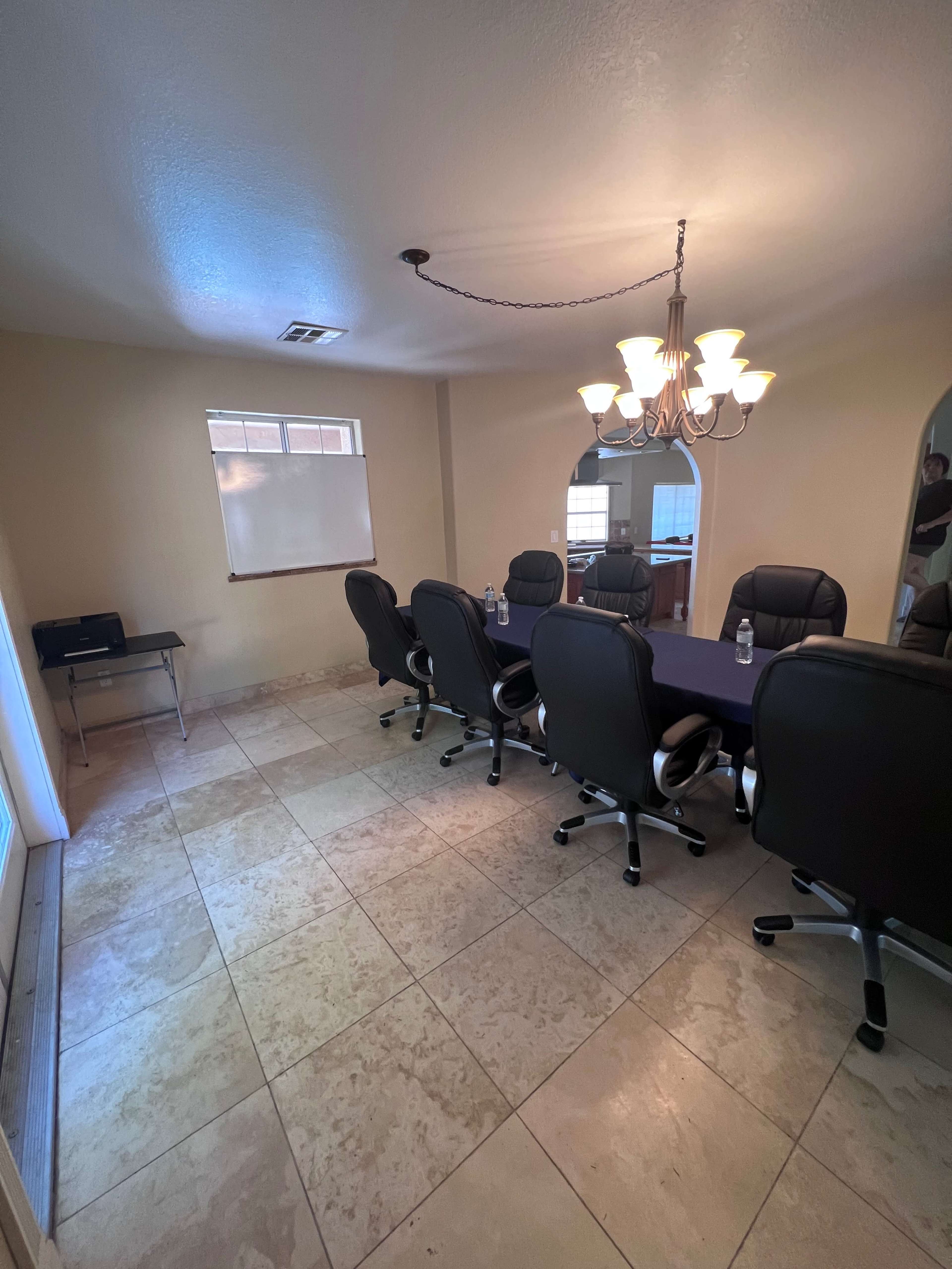 A conference room with a long table surrounded by black office chairs, featuring a chandelier and a window.