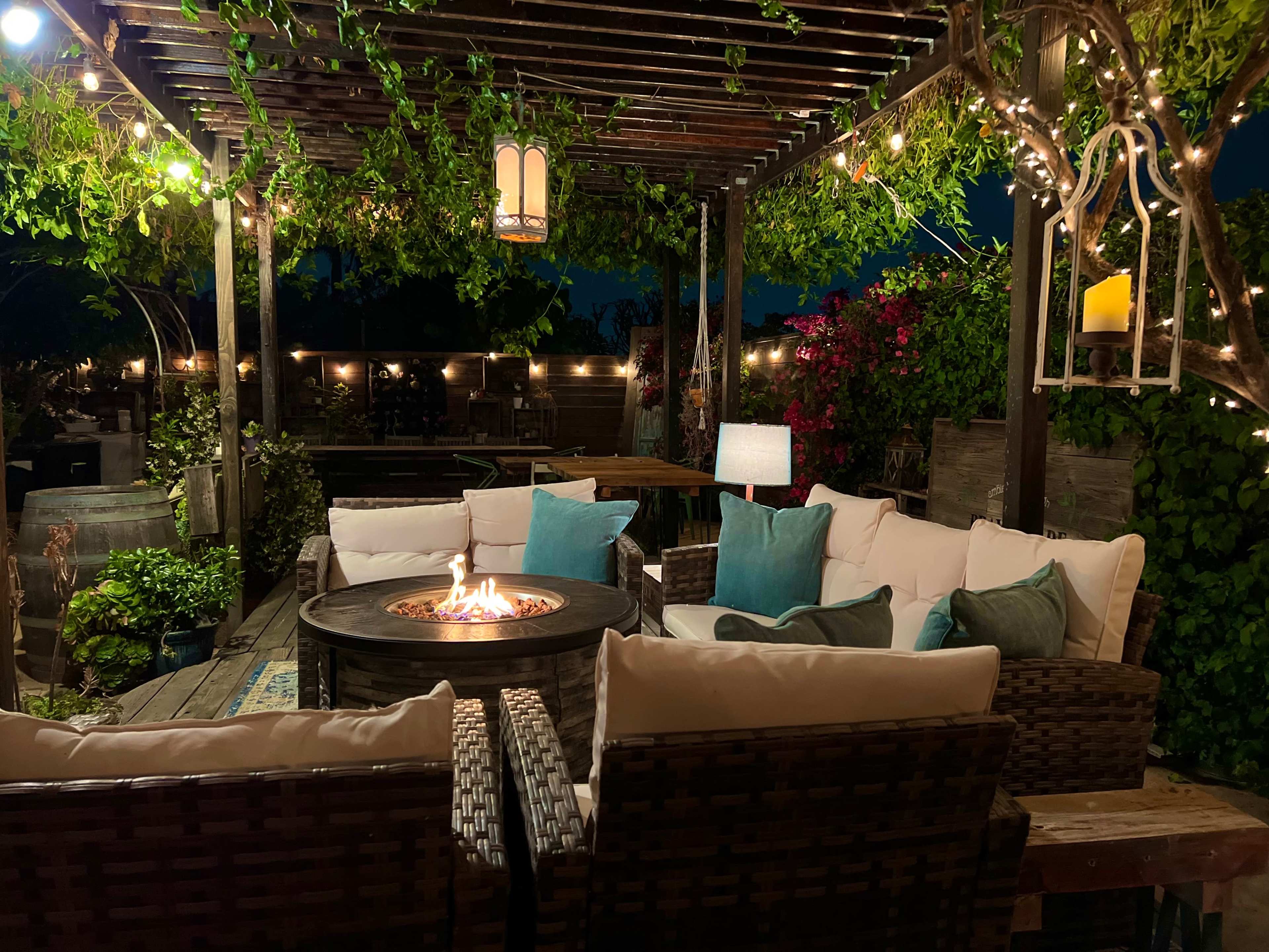 A cozy outdoor seating area features a circular fire pit surrounded by wicker chairs with light blue cushions, illuminated by string lights and lanterns.