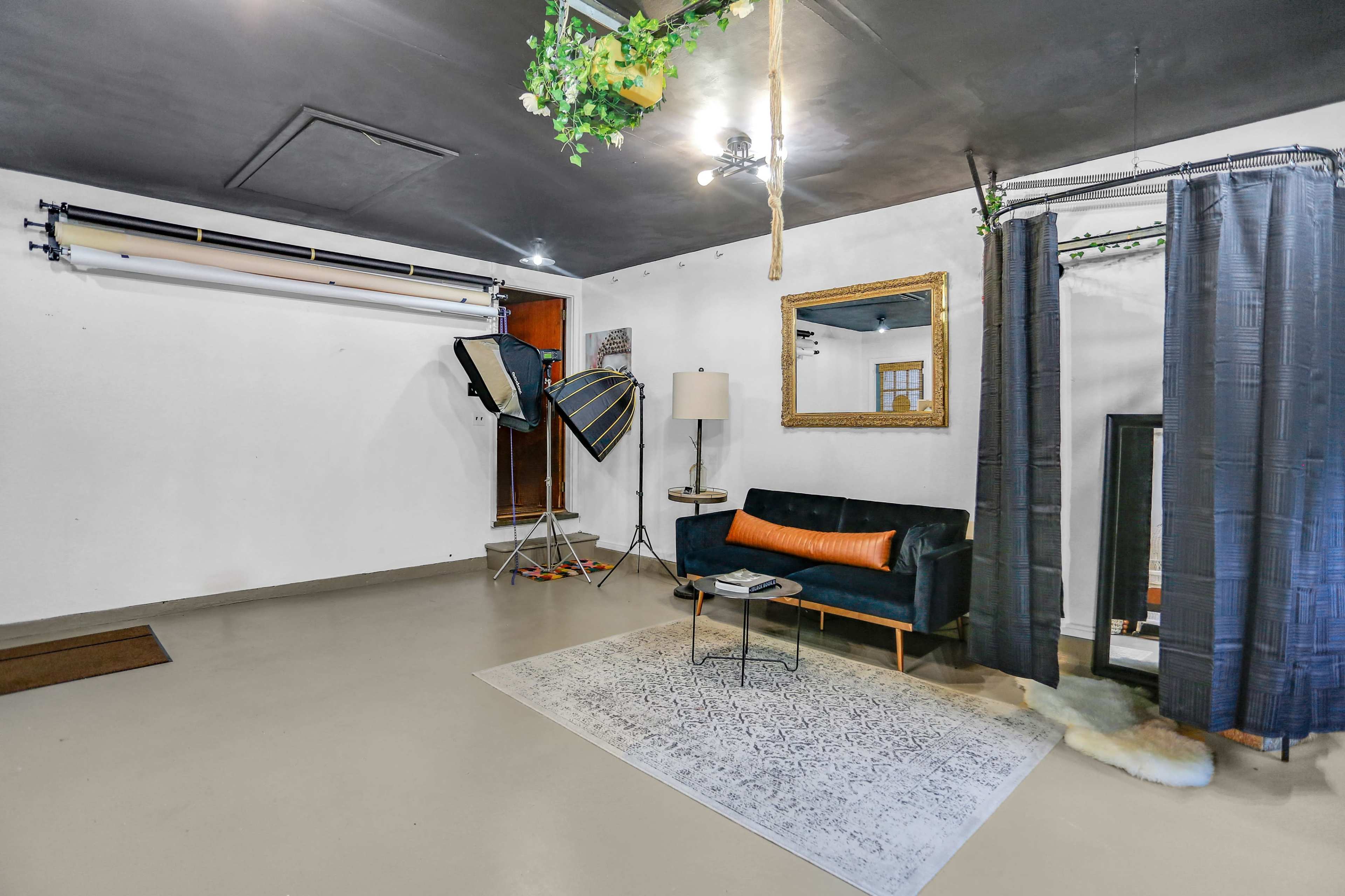 A spacious room with a black sofa, a standing lamp, a large mirror, a patterned rug, and photography equipment including softboxes and backdrops.