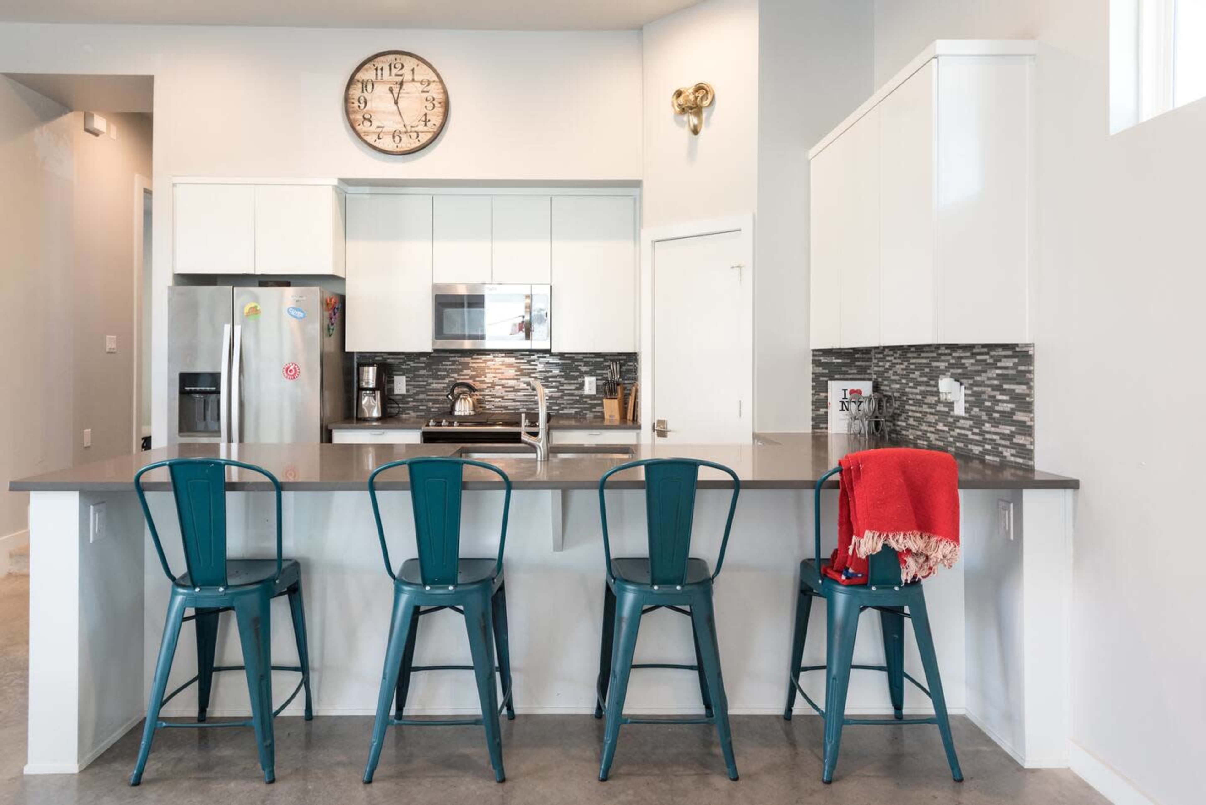 A modern kitchen features a gray countertop, four teal bar stools, stainless steel appliances, and a wall clock above the counter.