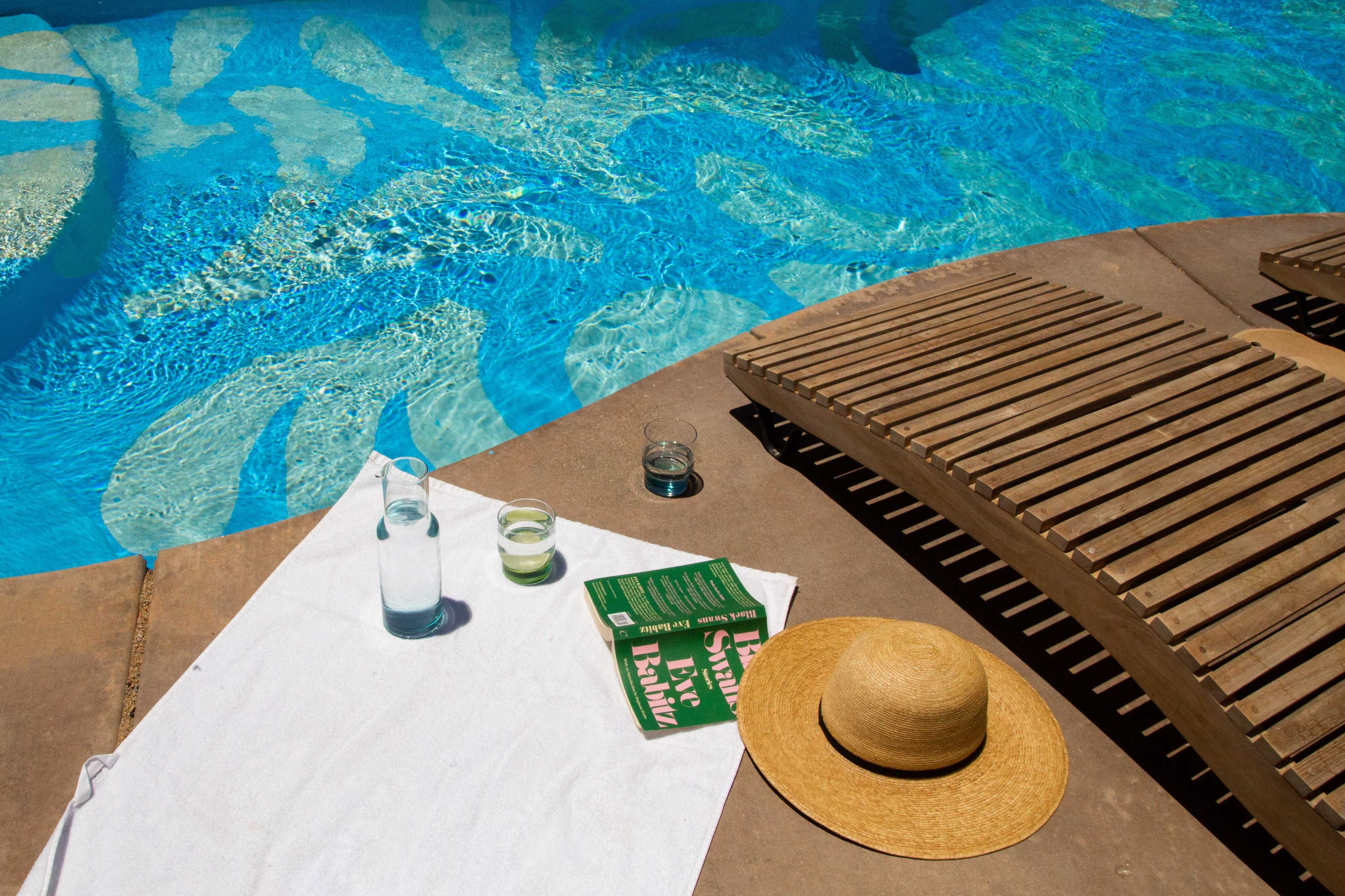 A poolside scene features a white towel, a straw hat, a book, and glasses of beverages beside a blue water surface.