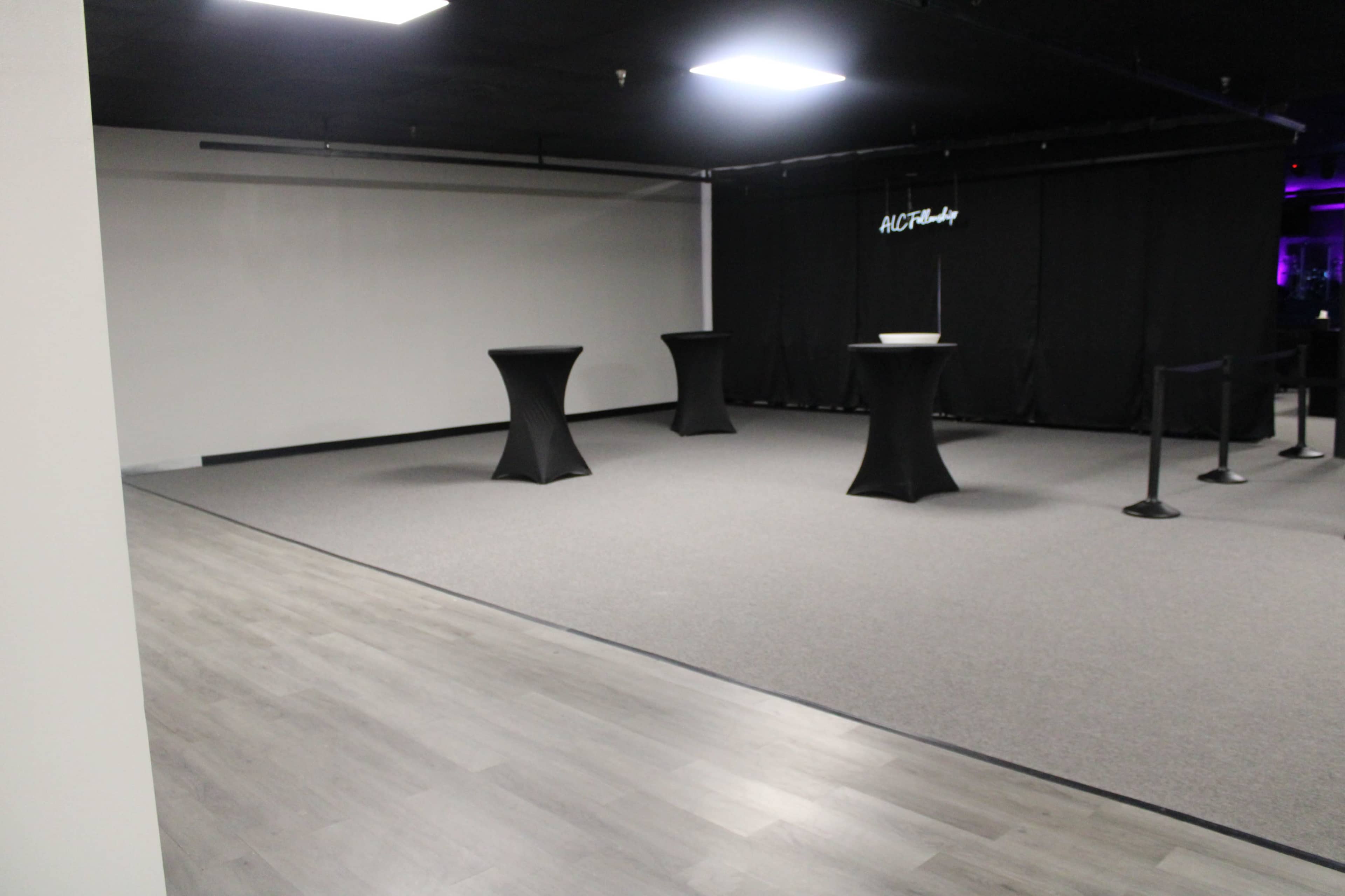 The image shows a spacious event venue with a gray carpet, featuring two round black tables and a black curtain in the background.
