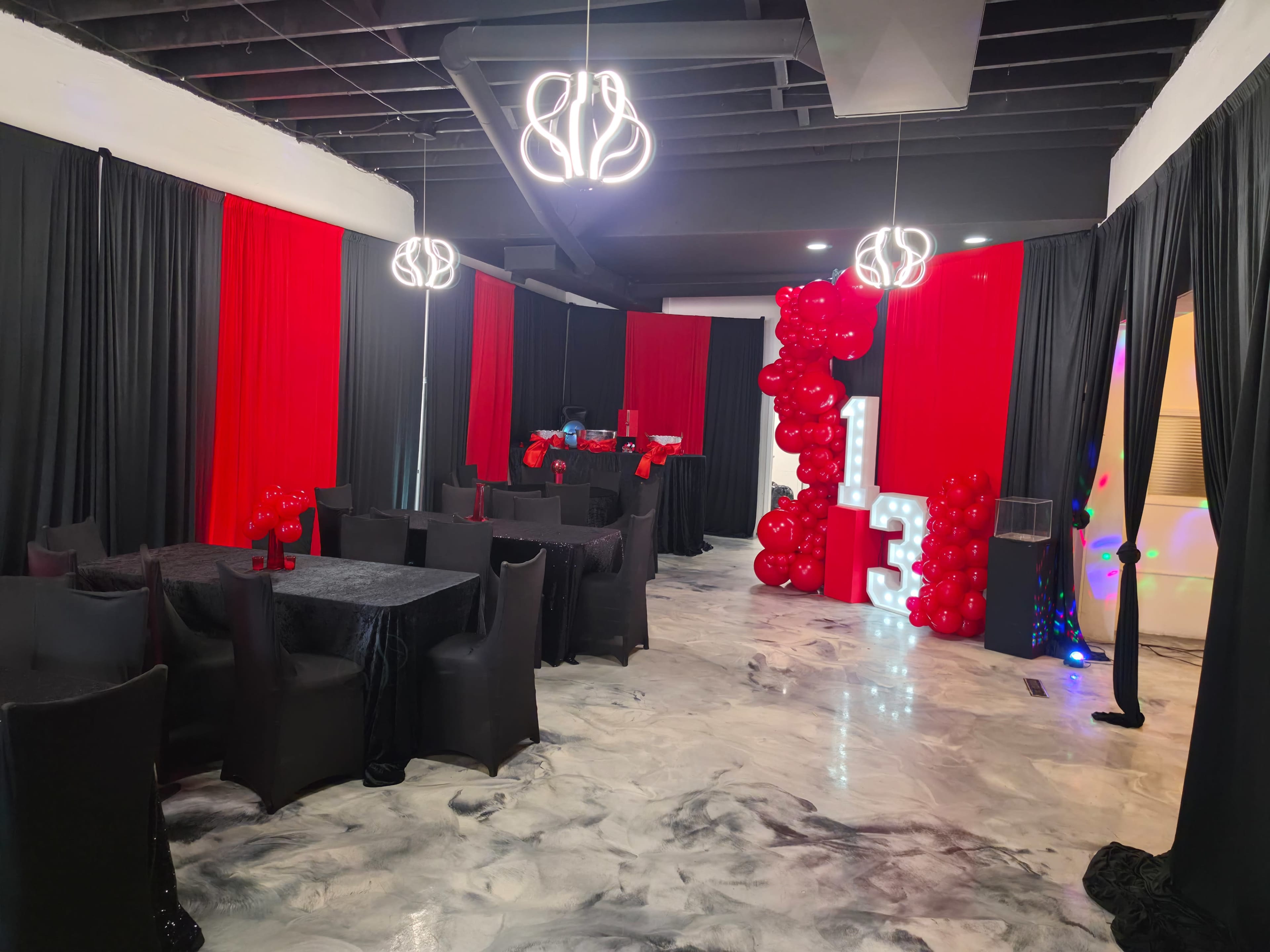 The image shows a decorated event space featuring black and red drapes, tables with black covers, and balloon arrangements spelling out the number "13."