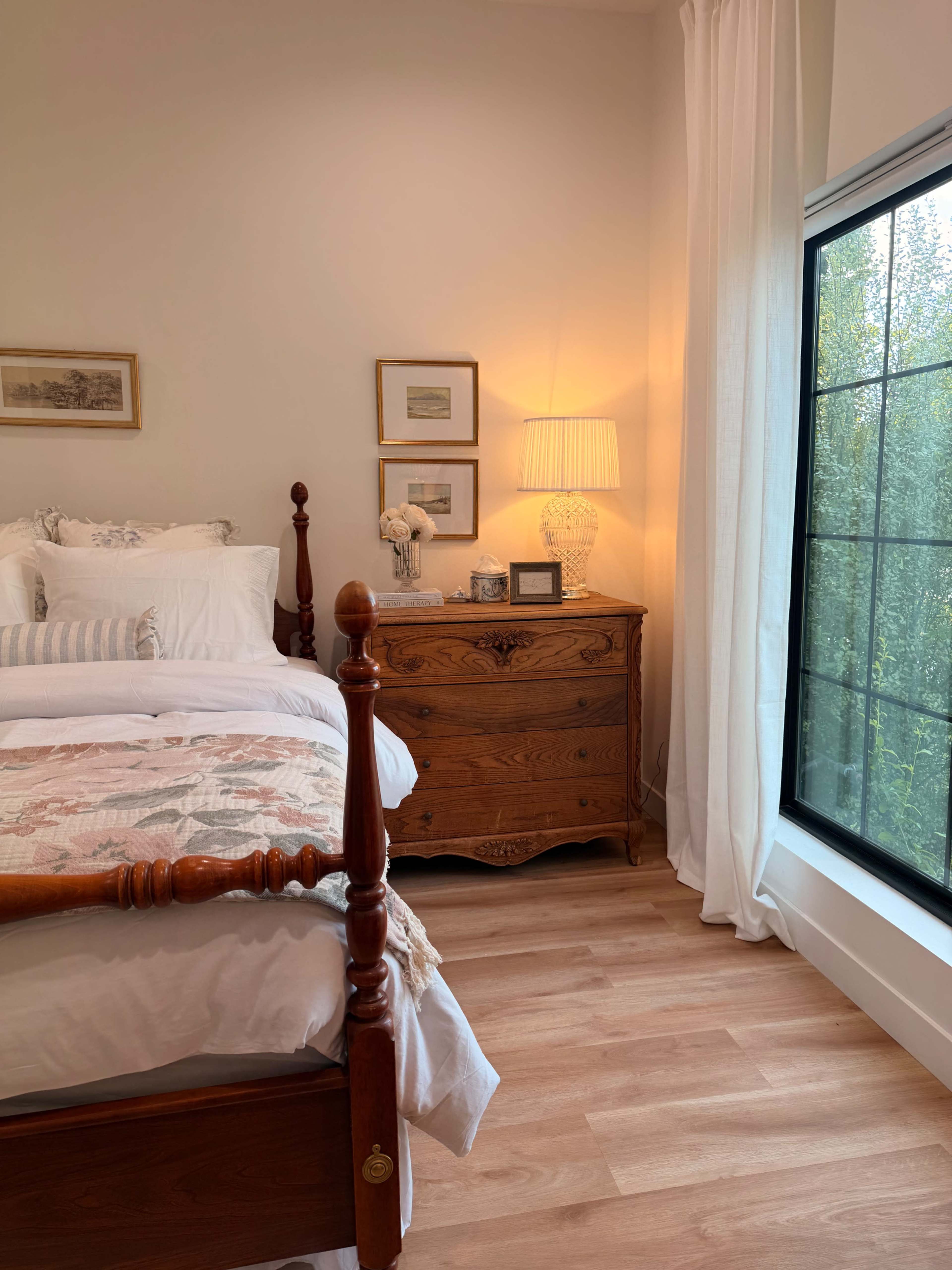 The image shows a bright bedroom with a wooden bed, a dresser, and a window featuring sheer curtains that reveal greenery outside.