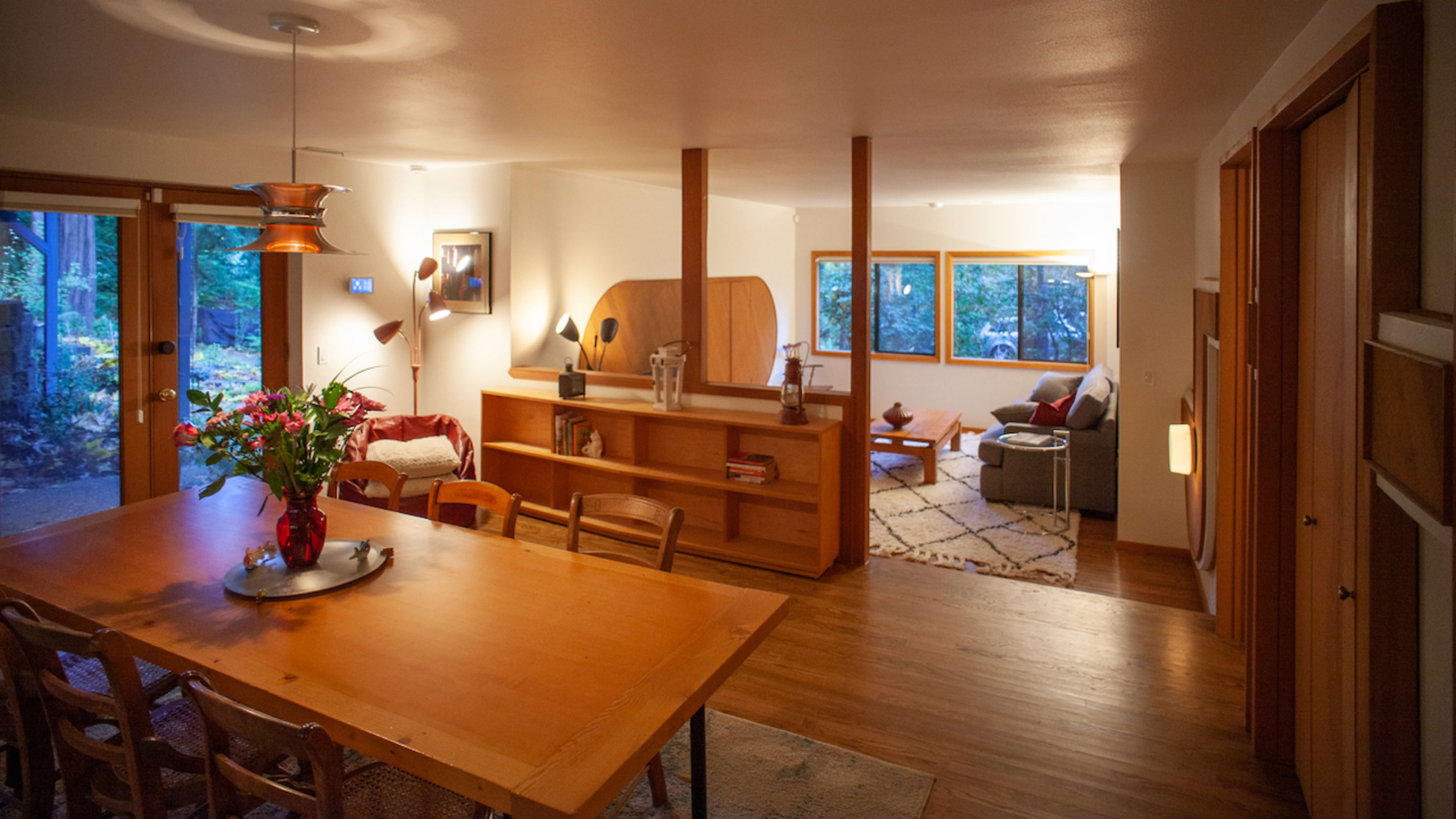 A wooden dining table is set in a well-lit open space, leading into a cozy living area with large windows overlooking a wooded view.