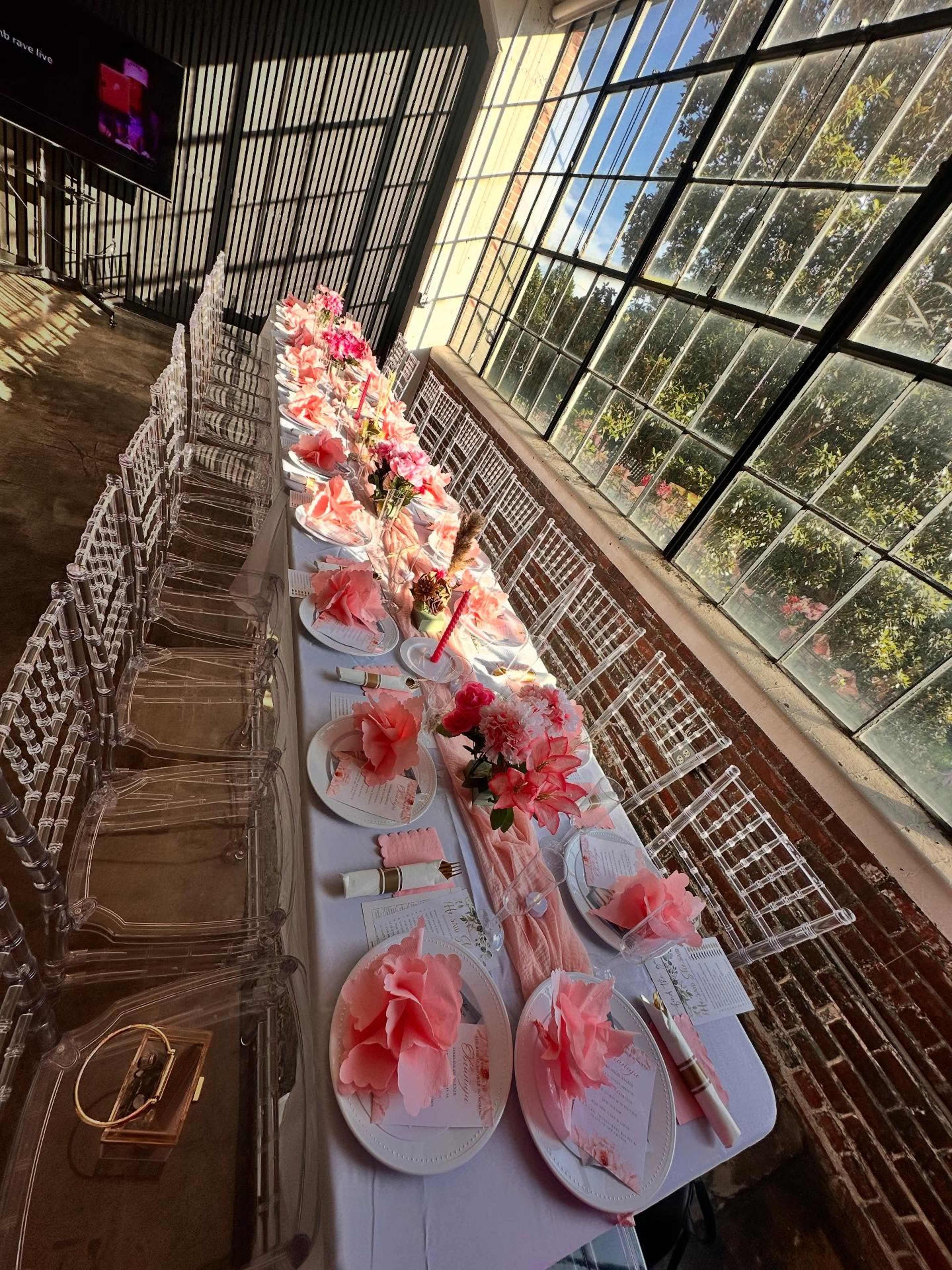 A long dining table is decorated with pink floral arrangements and placed against large windows that let in natural light.
