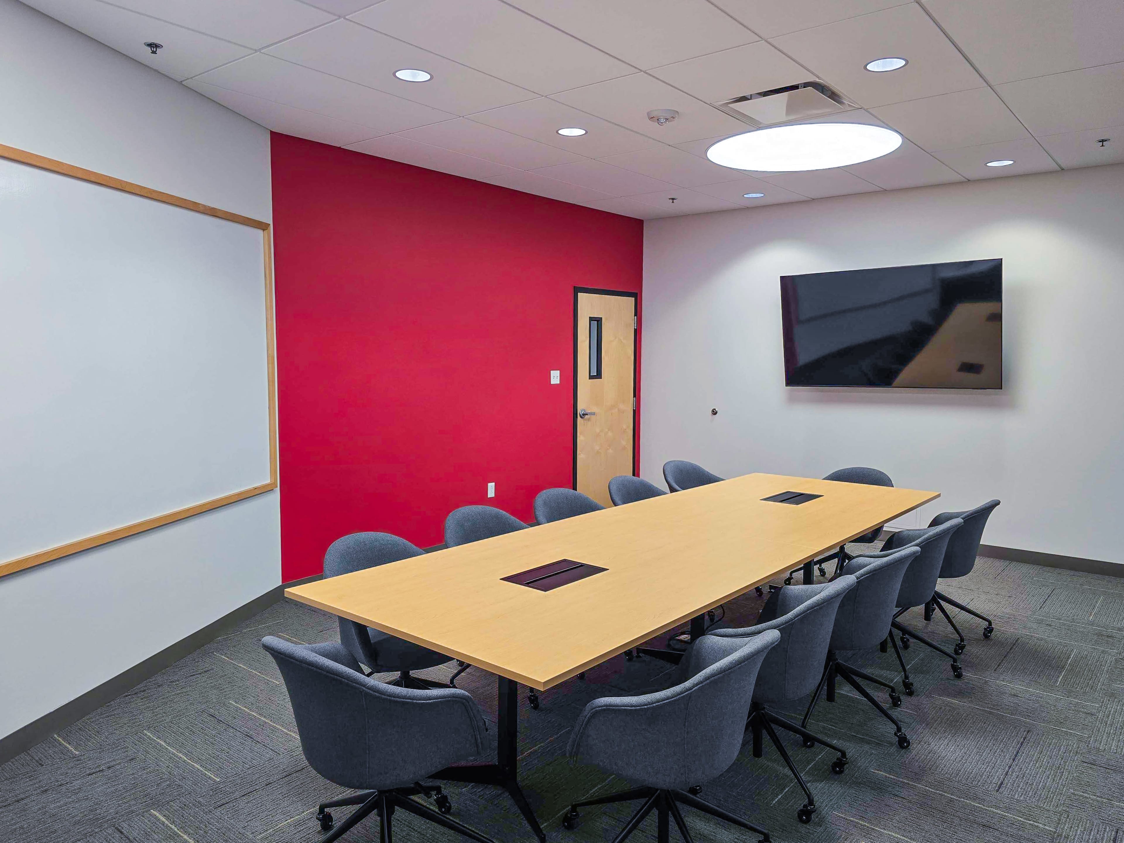 A conference room features a large wooden table surrounded by eight grey chairs, with a red accent wall, a whiteboard, and a mounted television.