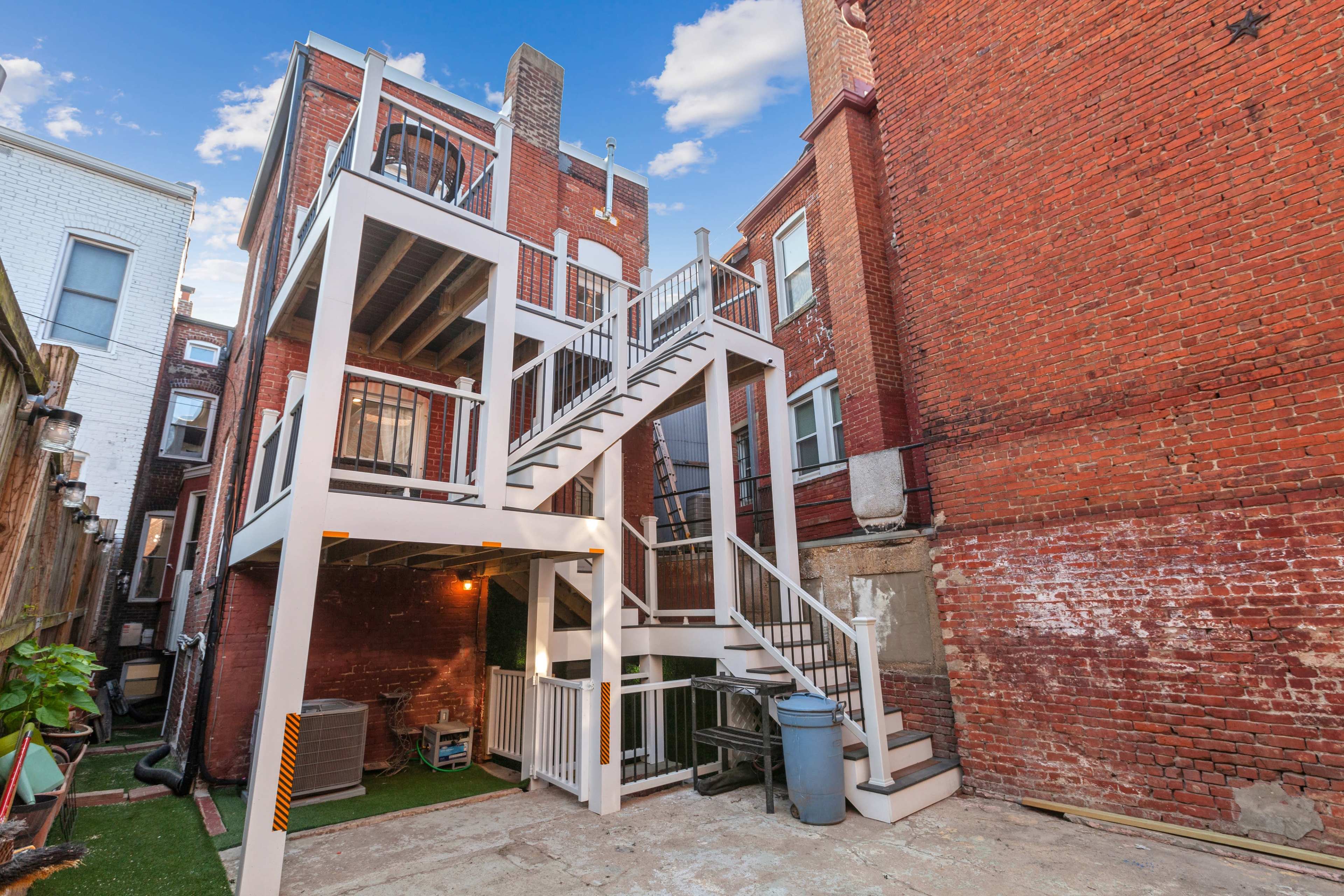 The image shows a two-story brick building with an exterior staircase leading to a balcony, situated in a narrow backyard area surrounded by other brick structures.