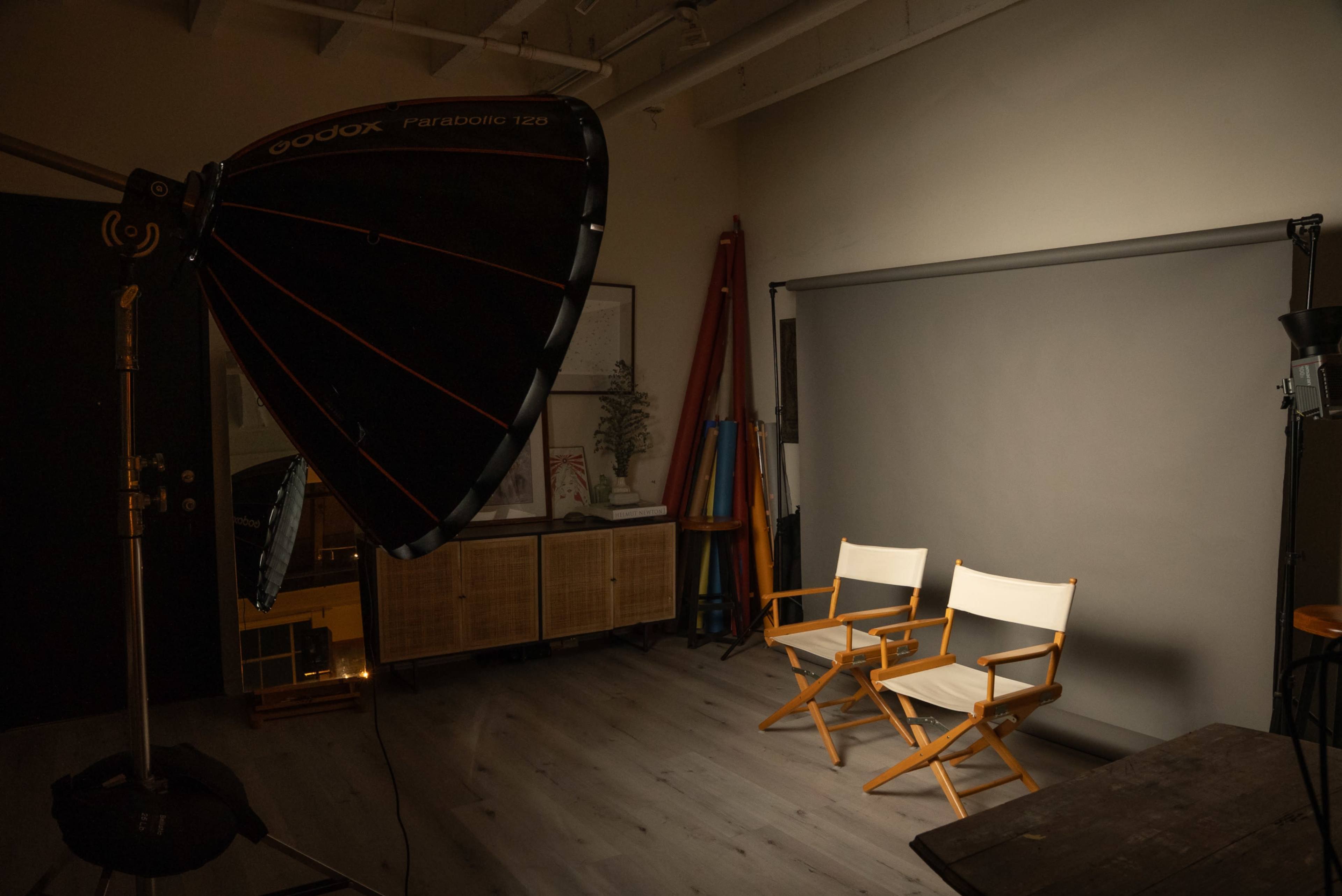The image shows a photography studio with two empty director's chairs positioned in front of a gray backdrop, illuminated by a large softbox light.