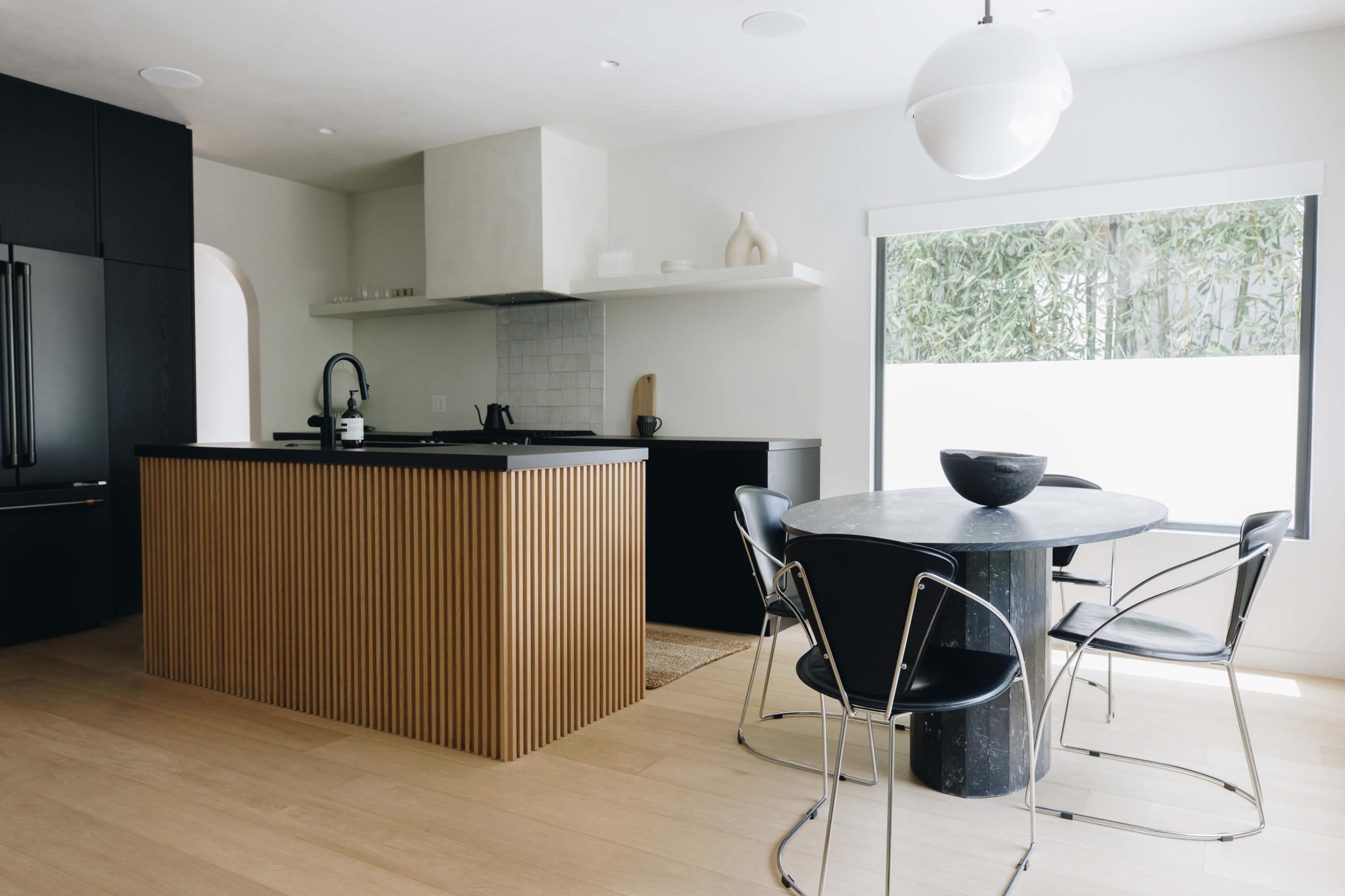 A modern kitchen features a black cabinet, a wooden island with vertical slats, and a round dining table with black chairs.