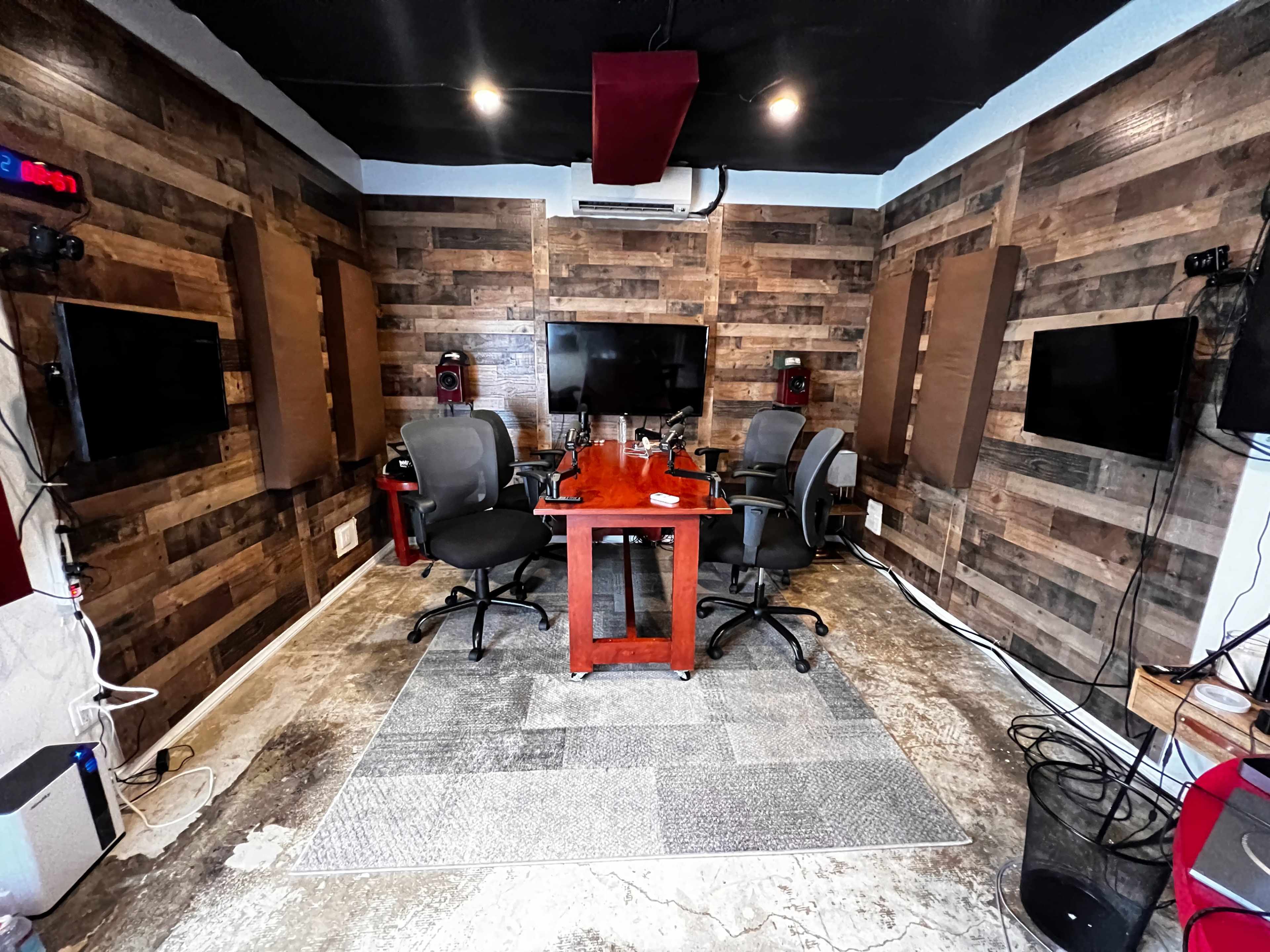 A small conference room features a wooden table surrounded by four chairs, with two wall-mounted screens and a large monitor against a wood-paneled backdrop.