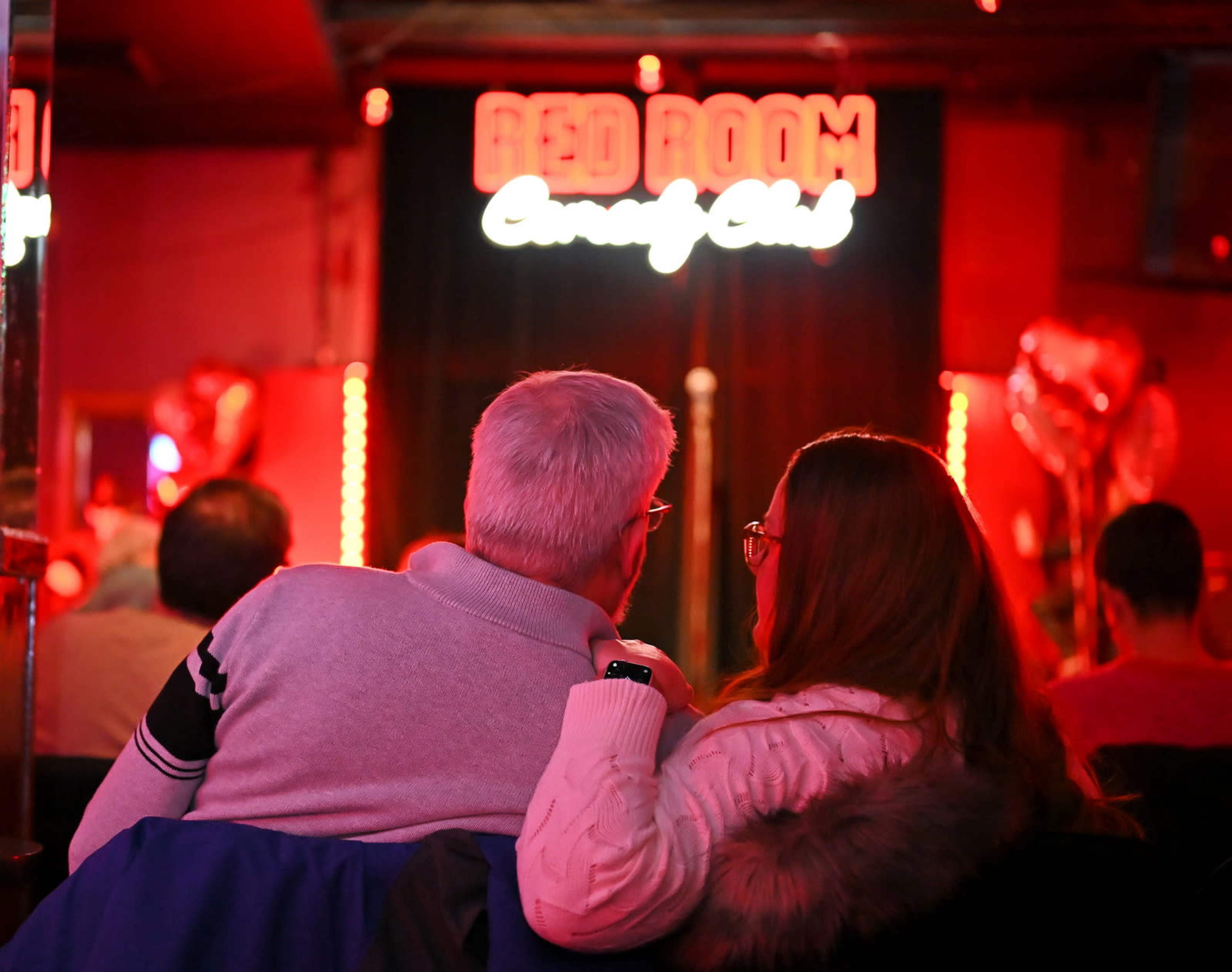 A couple sits closely together in a dimly lit comedy club, with a neon sign reading "Red Room Comedy Club" in the background.