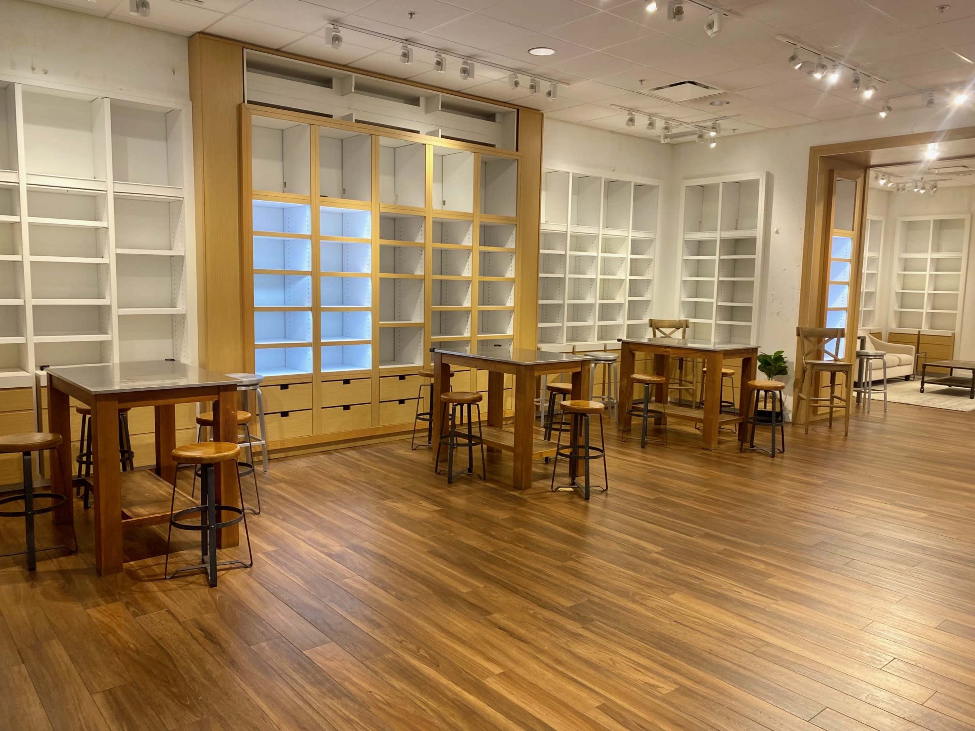 A spacious room with wooden flooring, featuring several tall tables surrounded by stools and empty shelves along the walls.