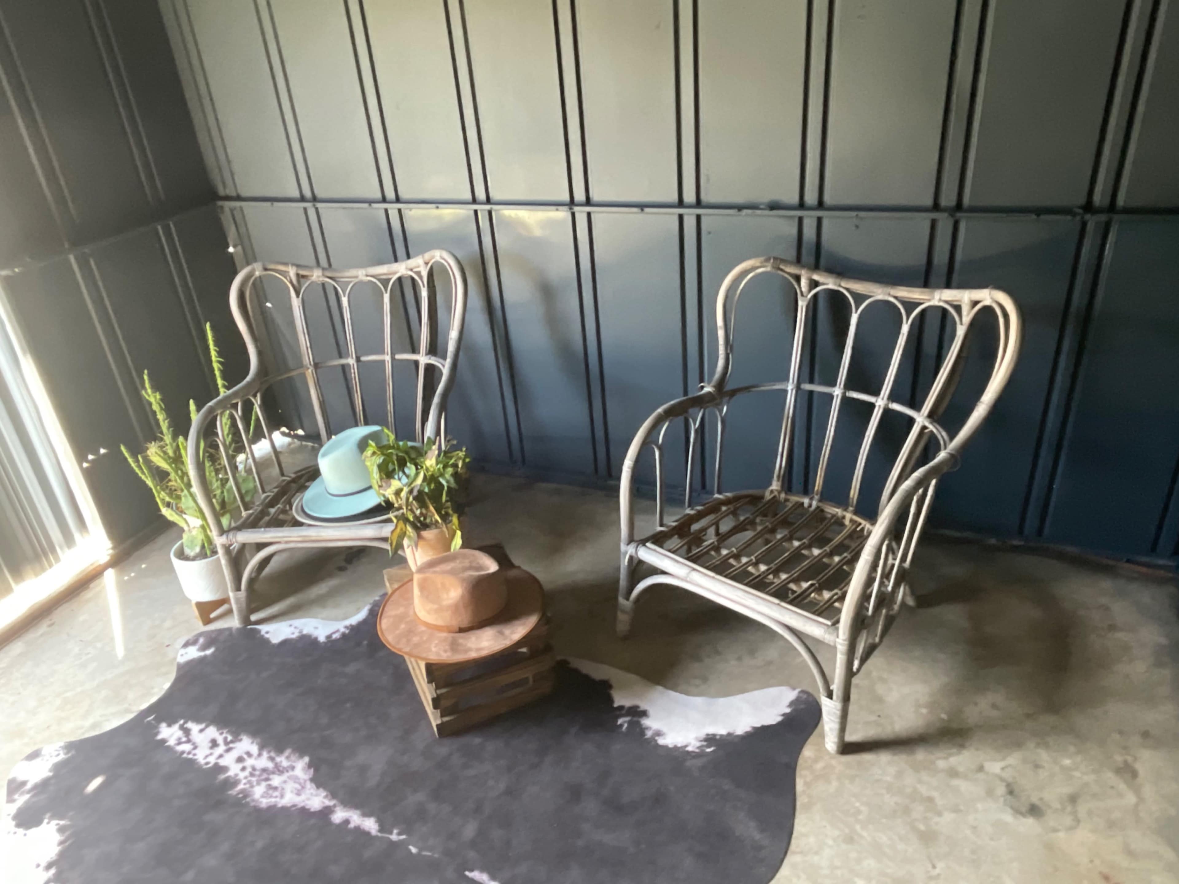 Two wicker chairs are positioned across from each other around a small, rustic table on a cowhide rug in a room with dark blue walls.