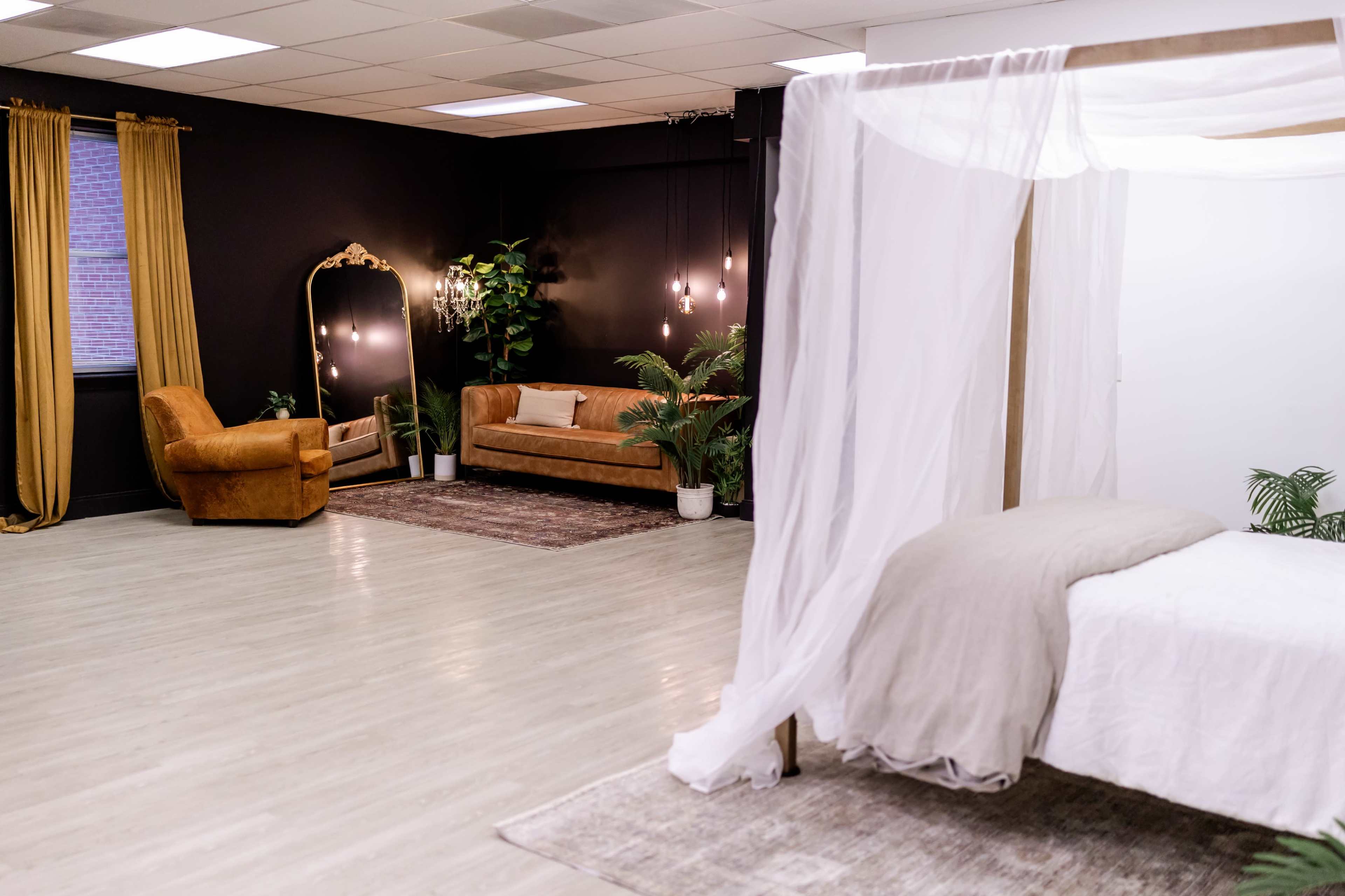 The image shows a modern, spacious interior with a bed draped in white fabric on the right and a seating area with a brown couch, a chair, and a large mirror on the left.