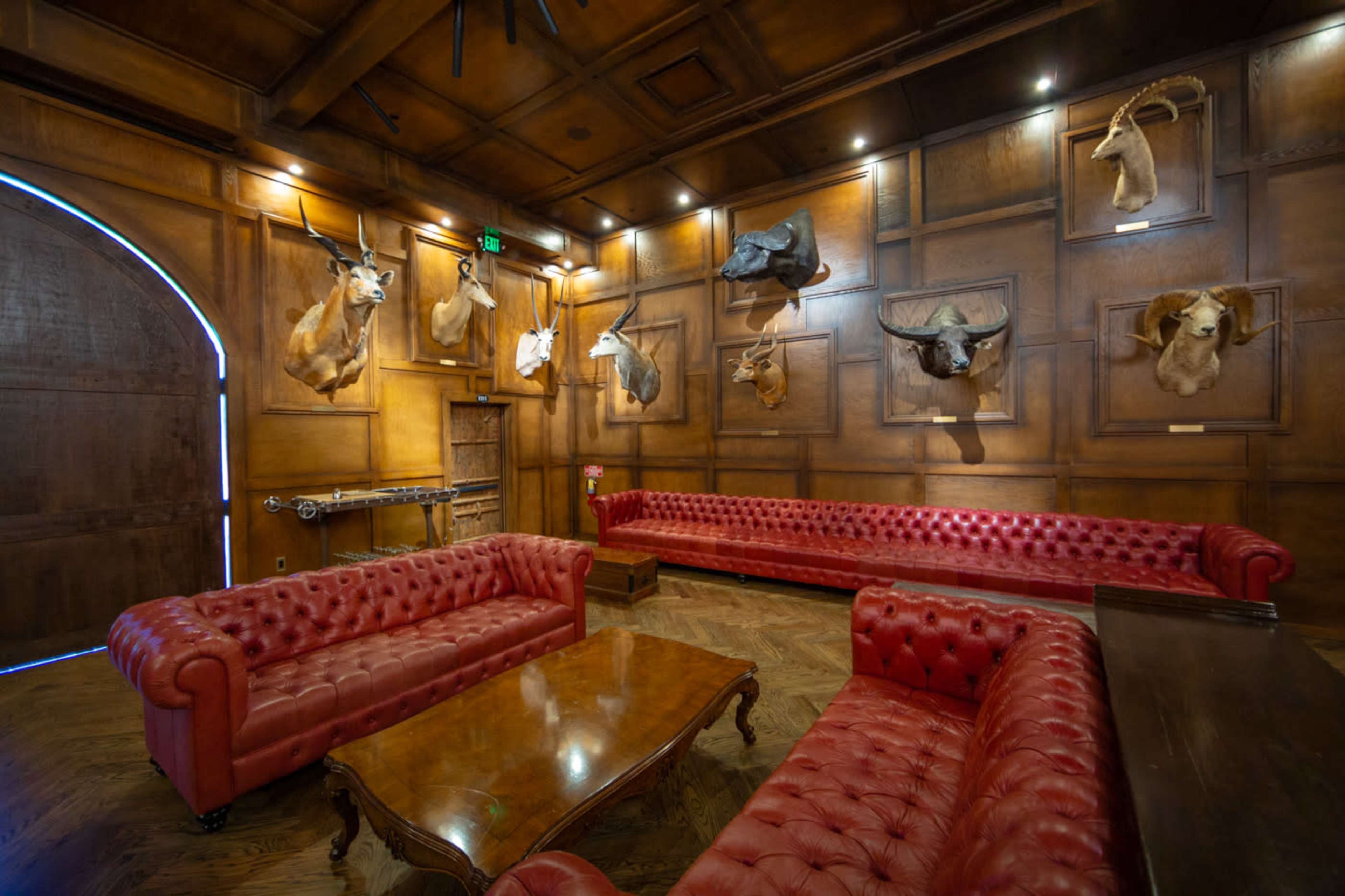 The image shows a room with wooden paneled walls, featuring multiple animal head mounts, and two red leather tufted sofas facing a wooden coffee table.