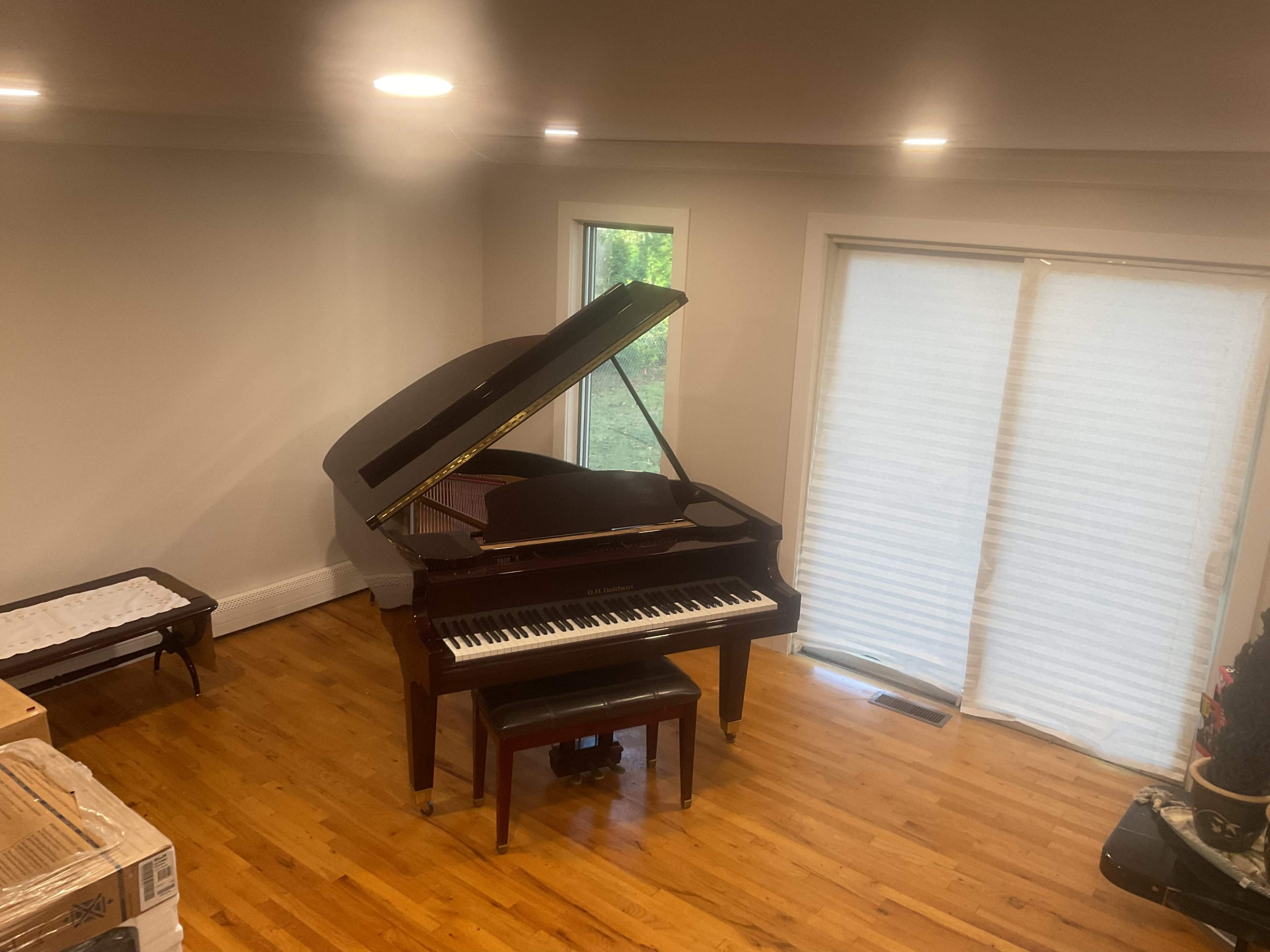 A grand piano with its lid raised is positioned in a well-lit room next to a large window, with boxes stacked nearby on the floor.