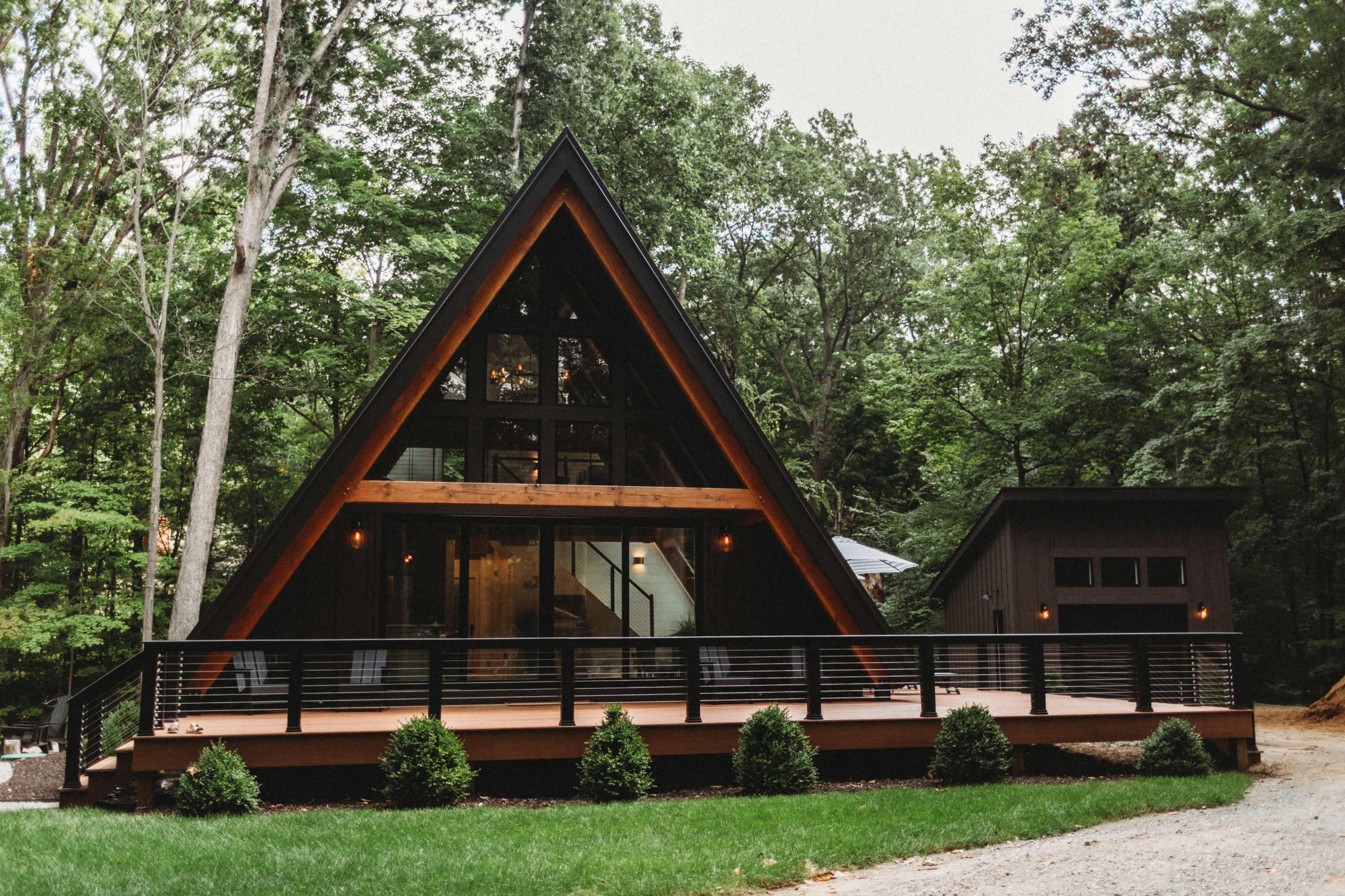 The image shows a modern A-frame cabin nestled among trees, featuring large glass windows and a wooden deck.