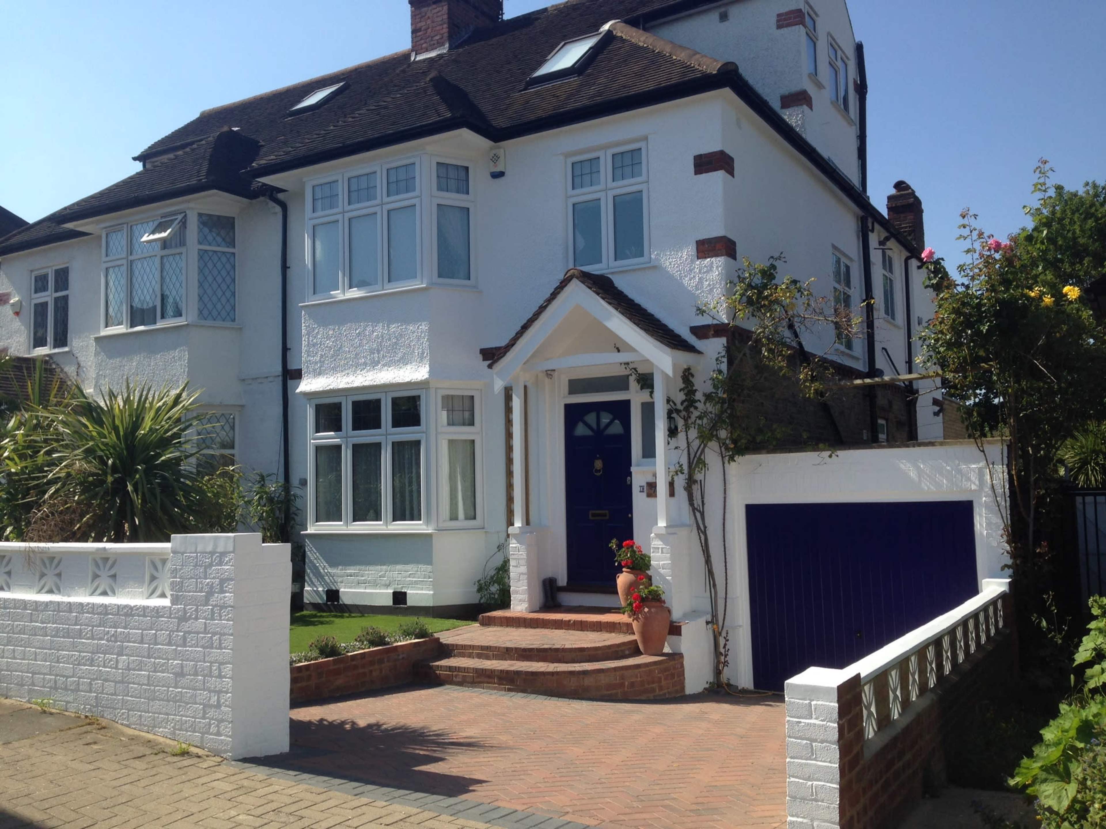 The image shows a two-story white house with a blue door and a brick driveway, surrounded by well-maintained gardens.