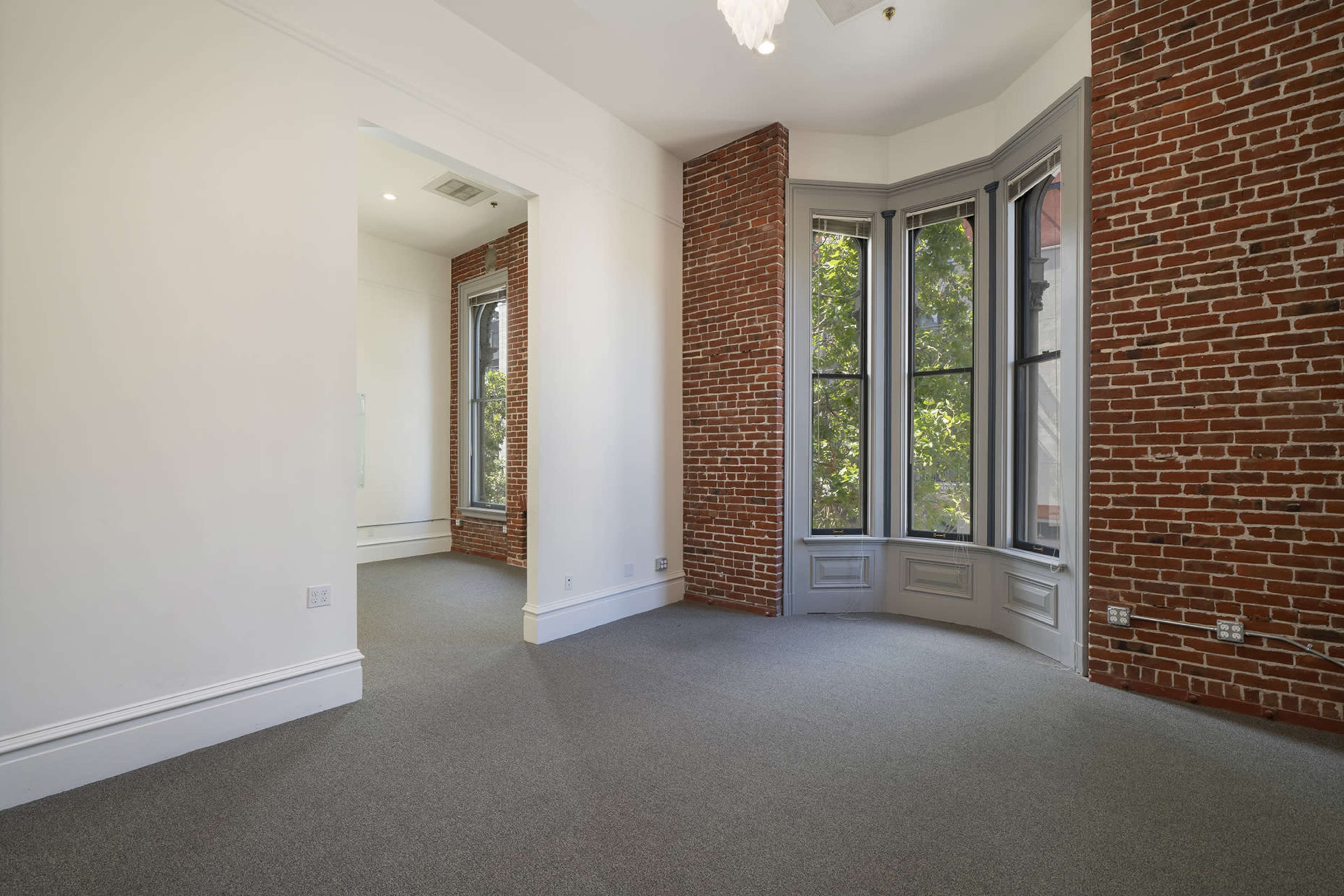 A spacious room with exposed brick walls, large windows, and a gray carpeted floor.