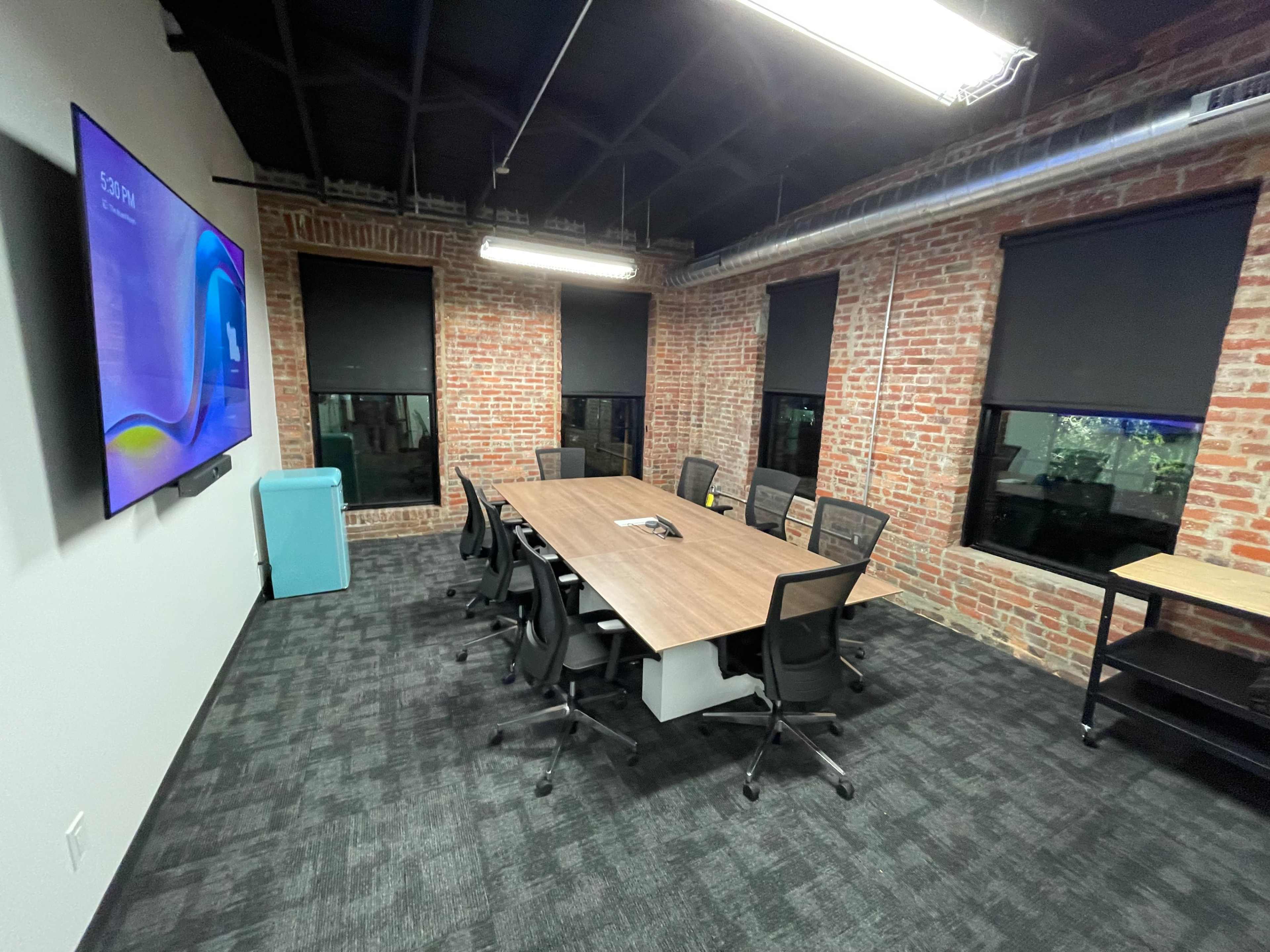 A conference room features a long wooden table surrounded by ergonomic chairs, with exposed brick walls and large windows, and is equipped with a wall-mounted screen.