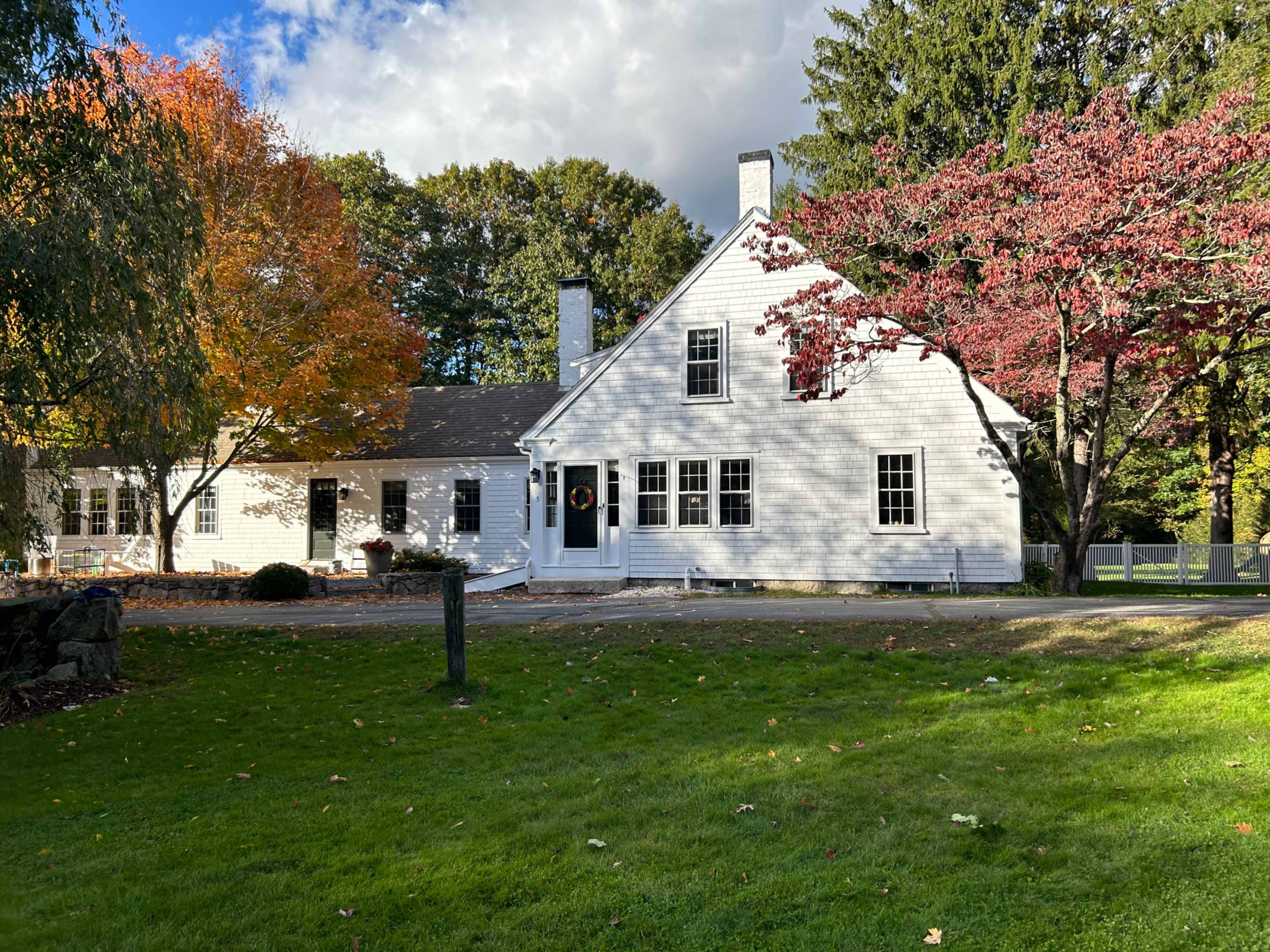 A white house with a gabled roof is surrounded by colorful autumn trees and a well-manicured lawn.