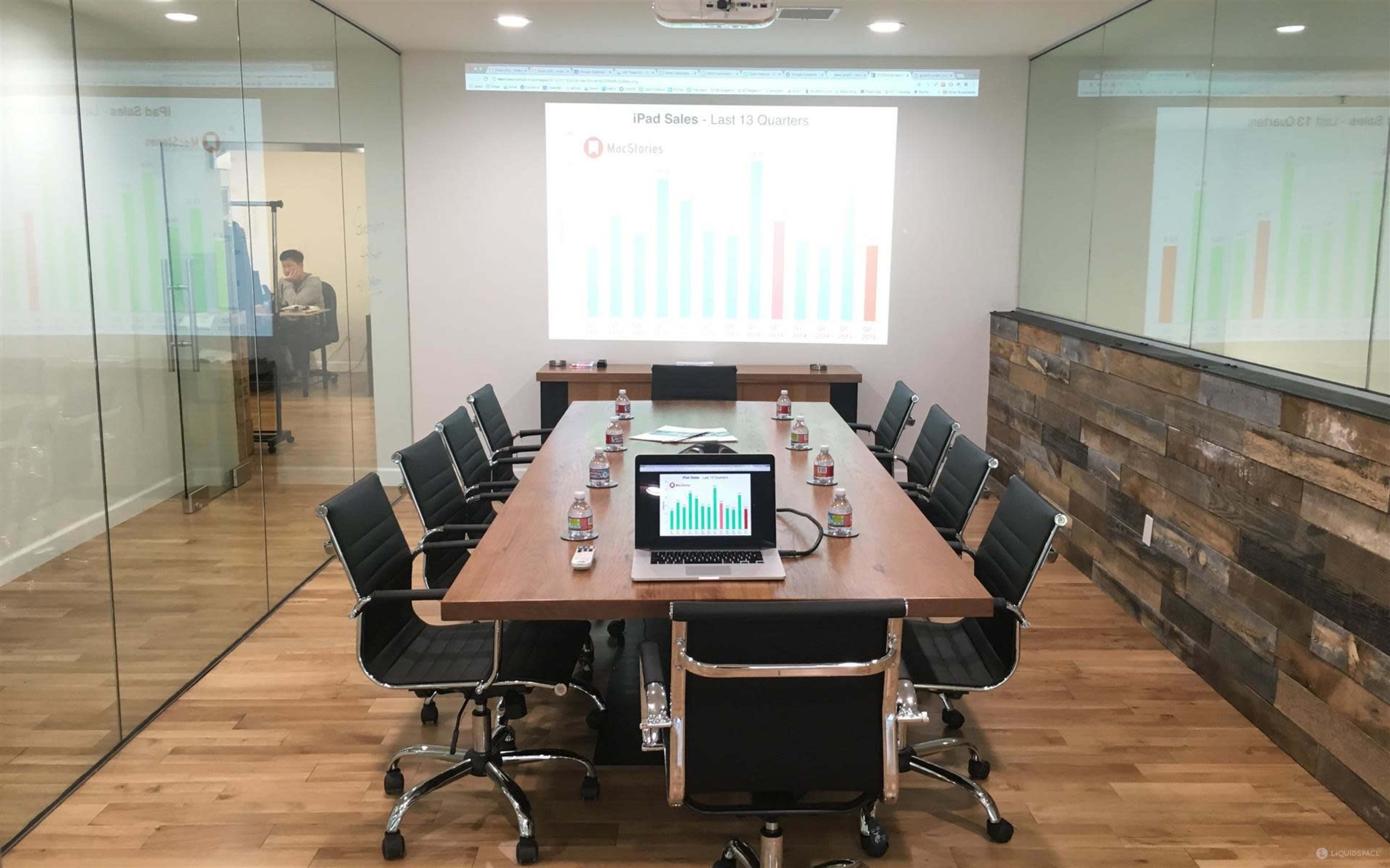 A conference room with a long wooden table, chairs, a laptop displaying a bar graph, and a presentation screen showing sales data, all set against glass walls.