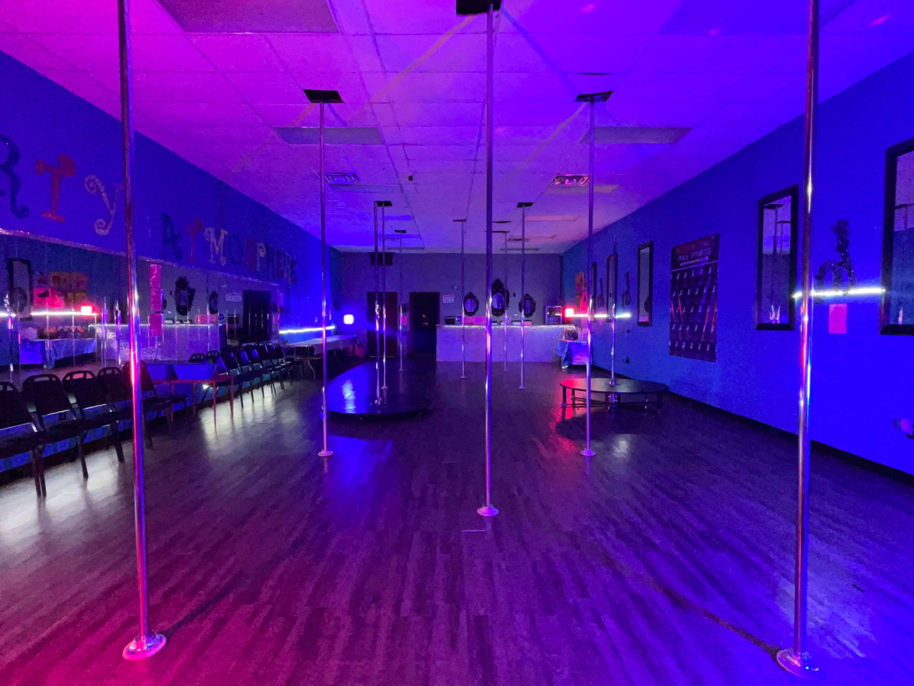 A dance studio with several pole dance poles, illuminated by purple and blue lights, and rows of chairs along one side.