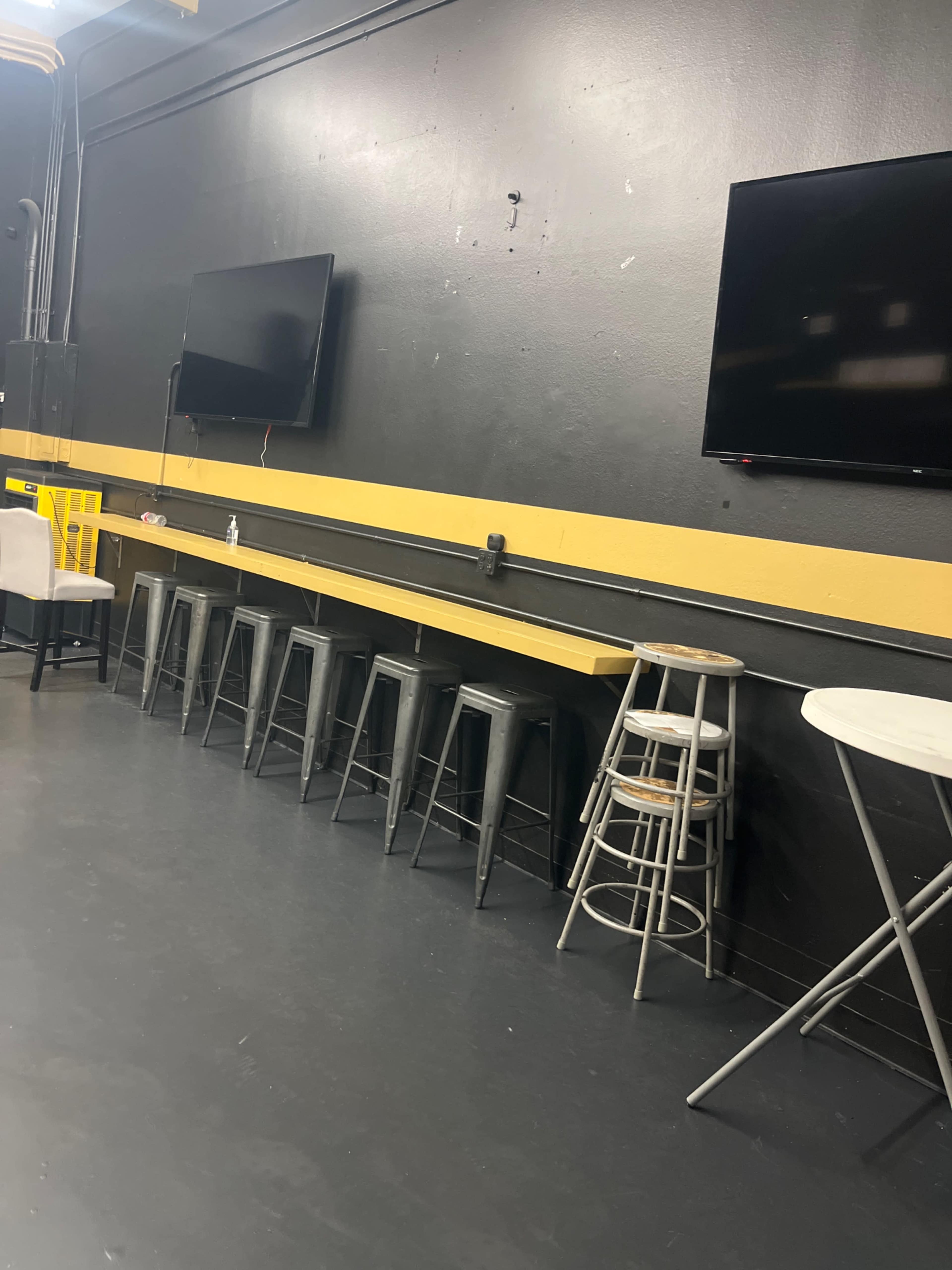 A black wall features two televisions, a yellow stripe, and several metal bar stools and tables arranged in a room.