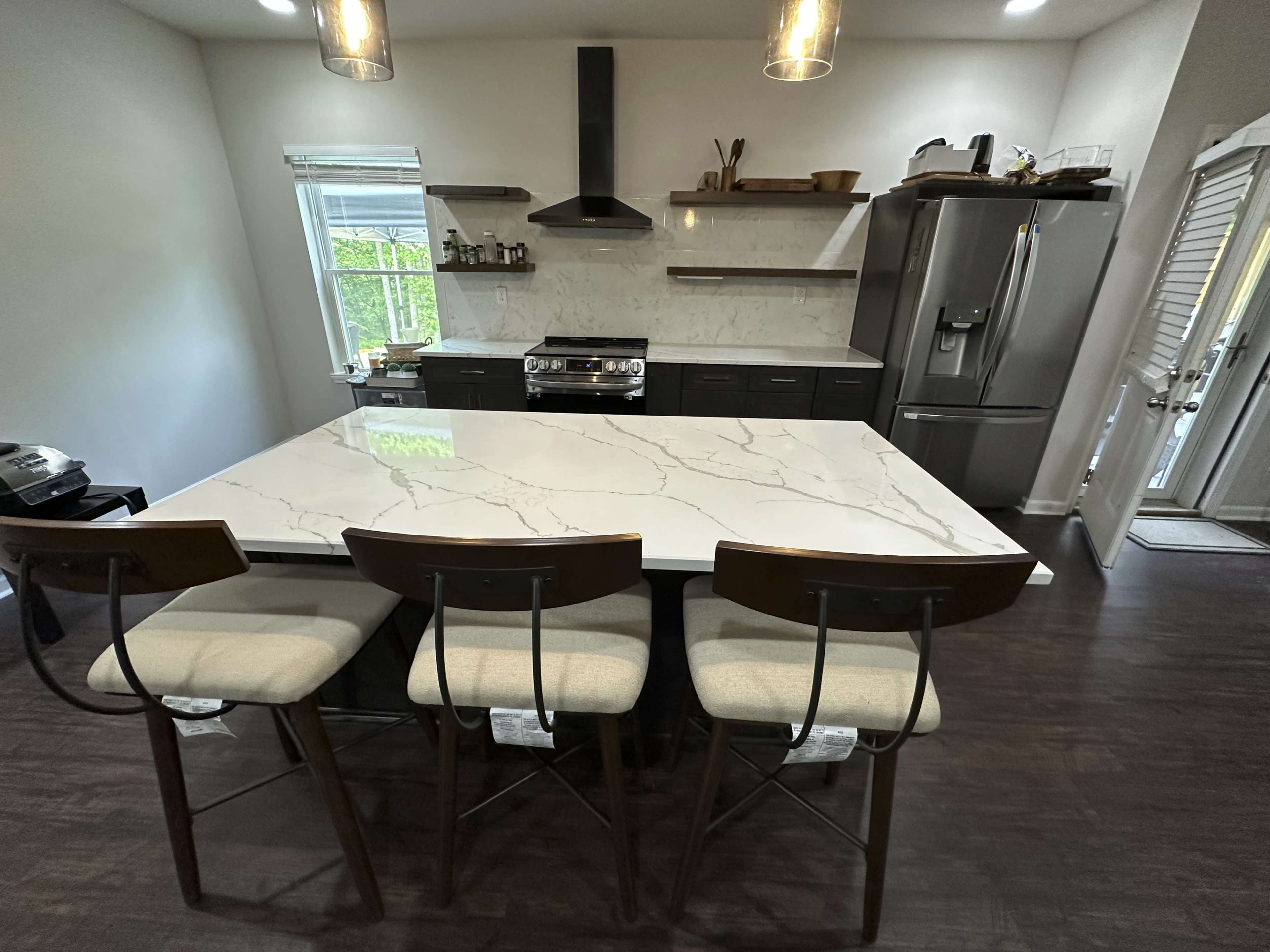 A modern kitchen features a large marble island with four wooden bar stools, stainless steel appliances, and minimalist cabinetry.