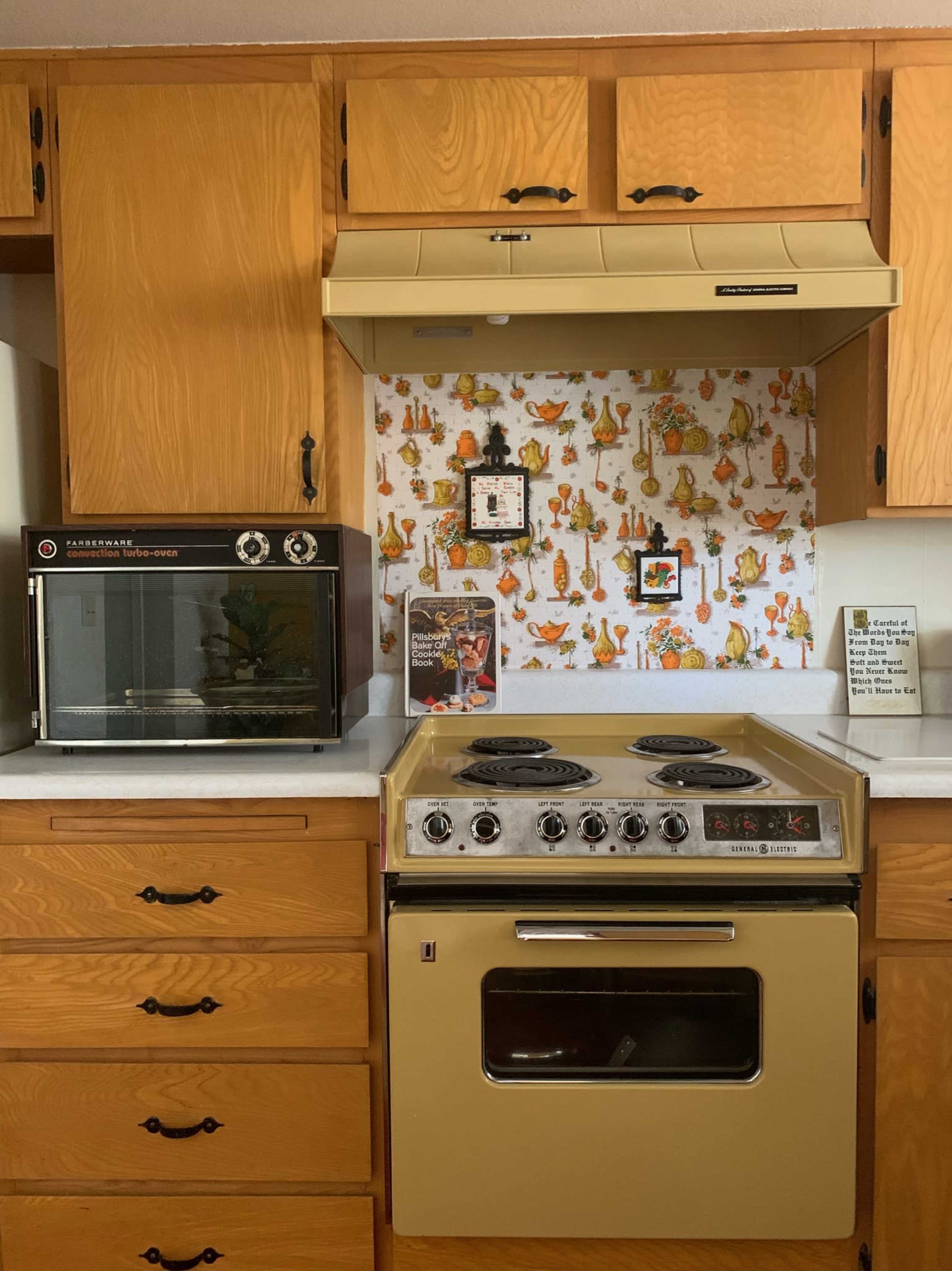 The image shows a kitchen with wooden cabinets, a vintage yellow stove, a countertop oven, and a patterned backsplash featuring various kitchenware.