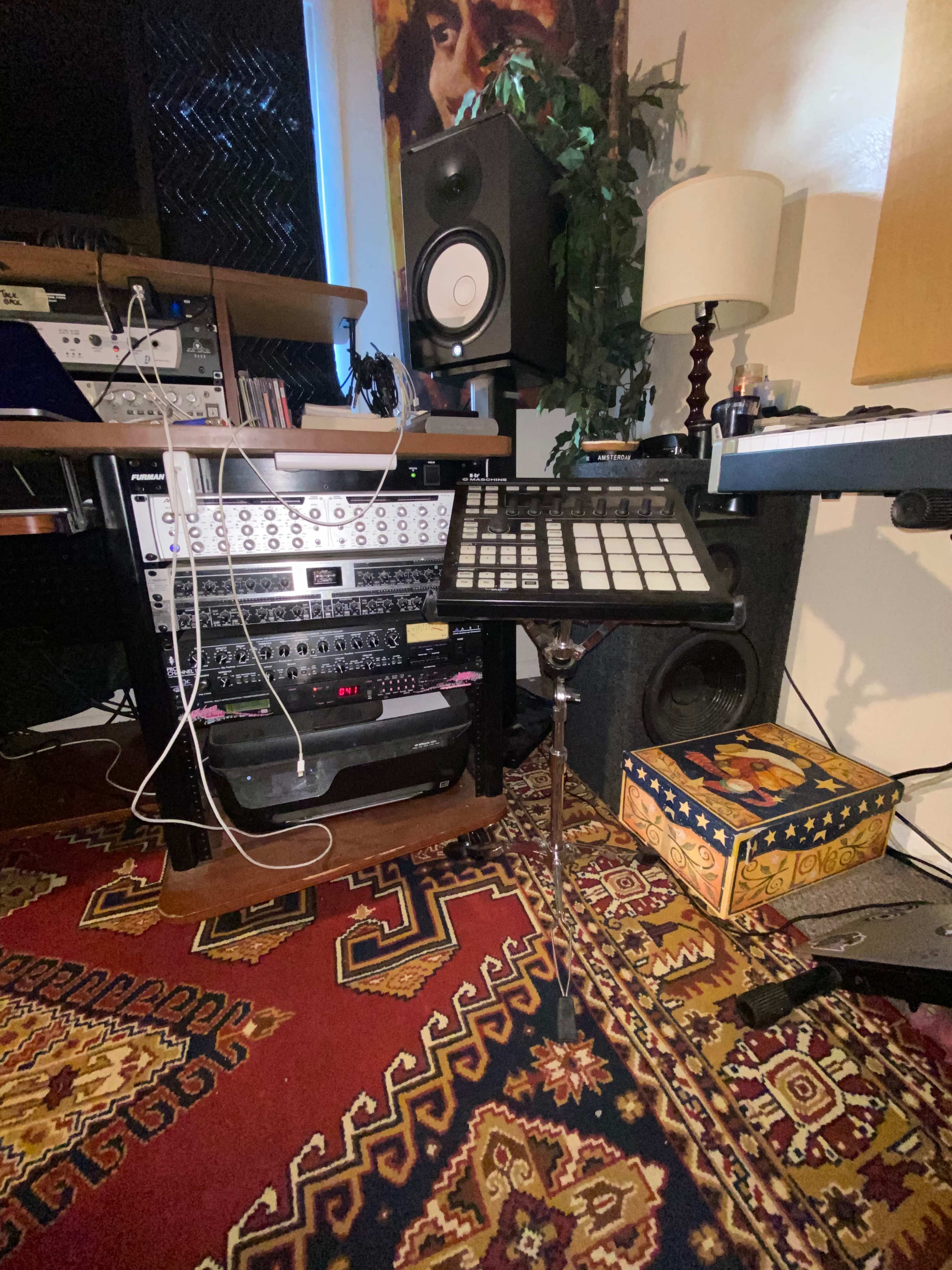 The image shows a music production setup featuring various audio equipment, a keyboard controller, a lamp, and decorative storage on a patterned rug.