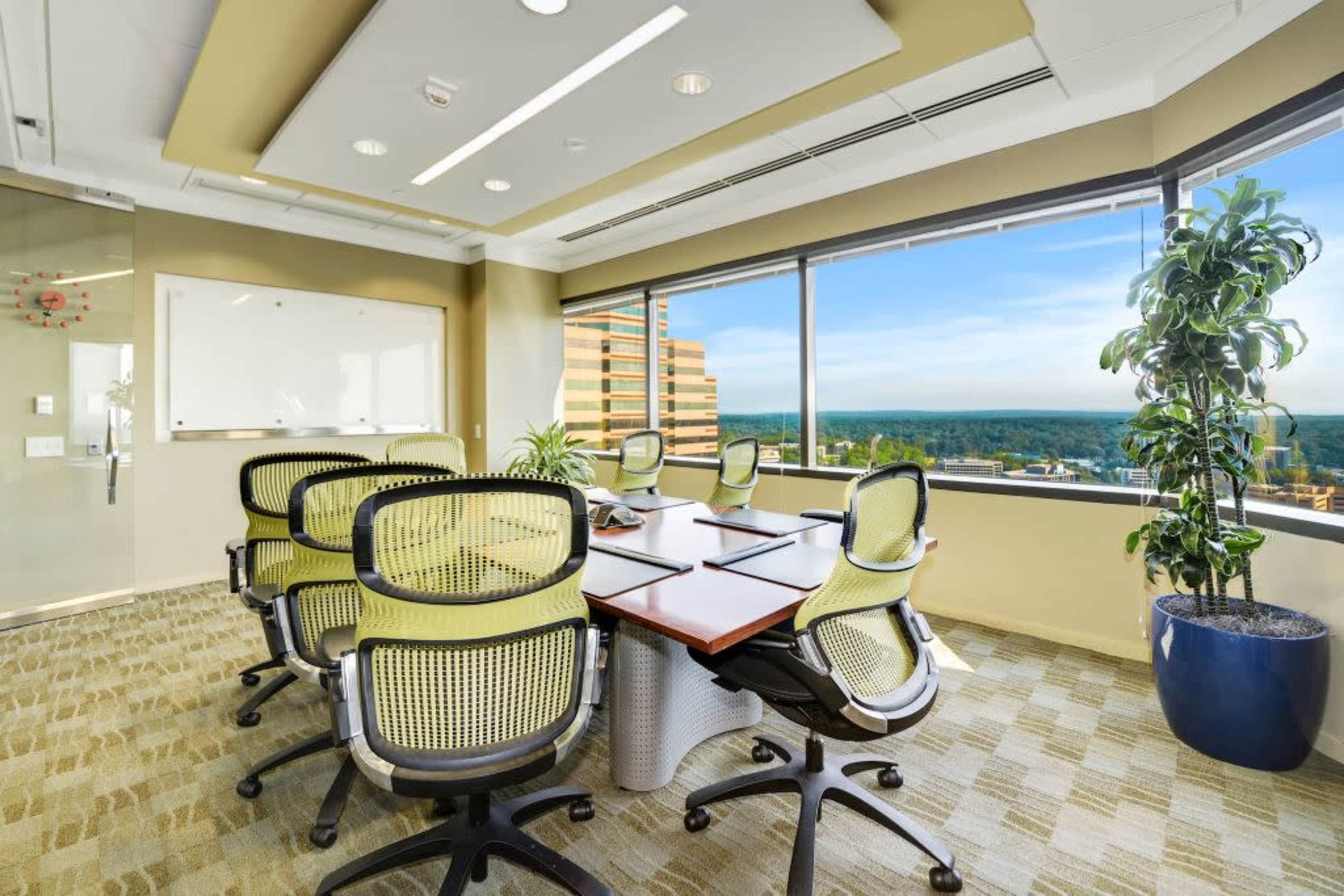 A modern conference room features a large, oval table surrounded by six green office chairs and large windows offering a view of the landscape outside.